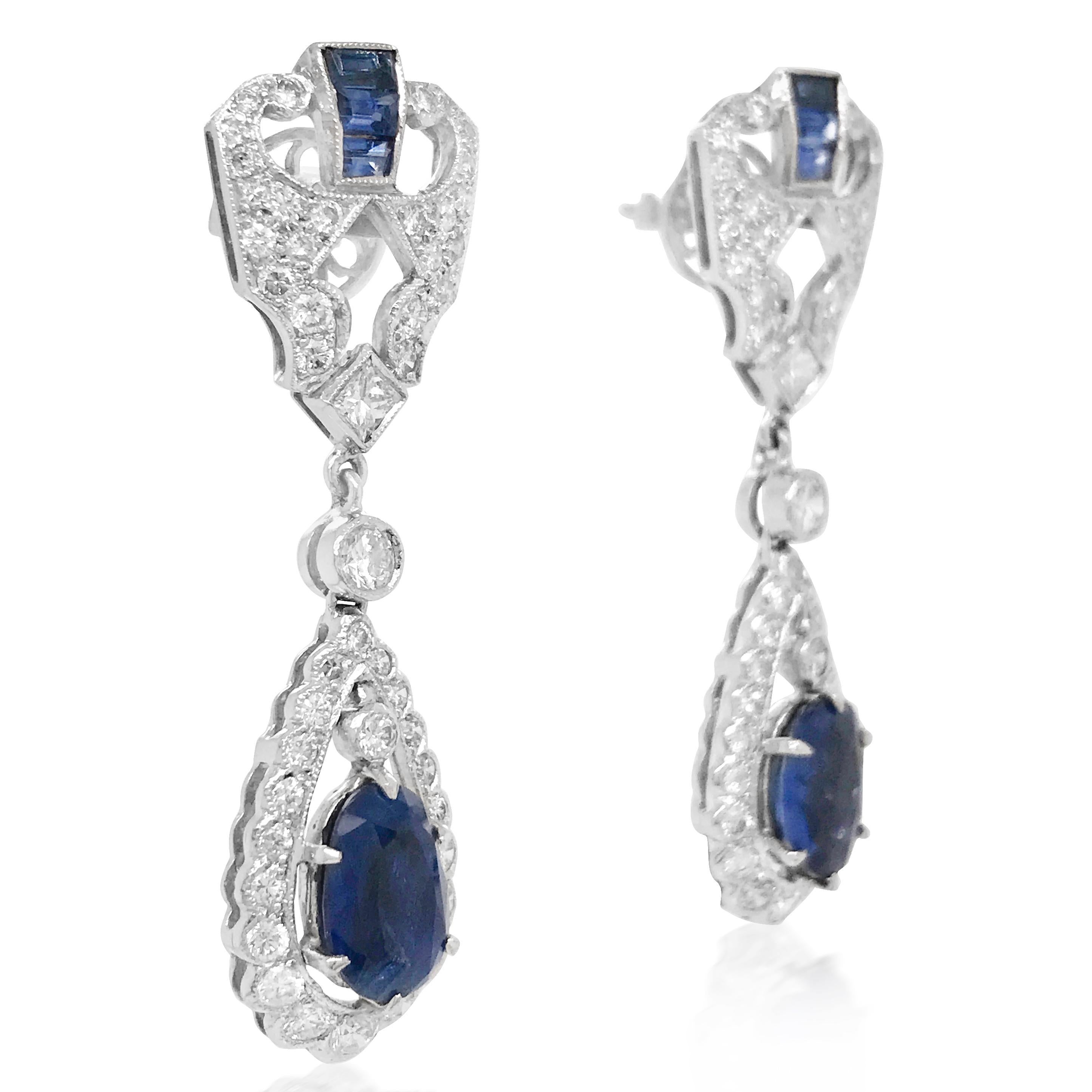 These stunning blue sapphire and diamond drop earrings are crafted in 18K white gold, weighing 10.26 grams and measuring 44mm long x 11mm wide. Exposing a pair oval-cut blue sapphires, each weighing approx 1.0ct. Topped with a pair of shield-shaped