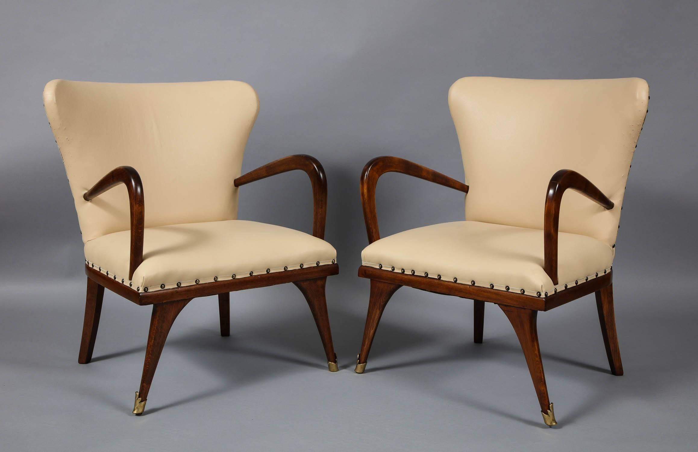 A pair of diminutive modernist armchairs in cream colored leather with studded seams having gently barreled backs, vaulting arms and swept front and back legs, the front legs with decorative brass sabots. (Upholstery as found), circa 1960s.

 