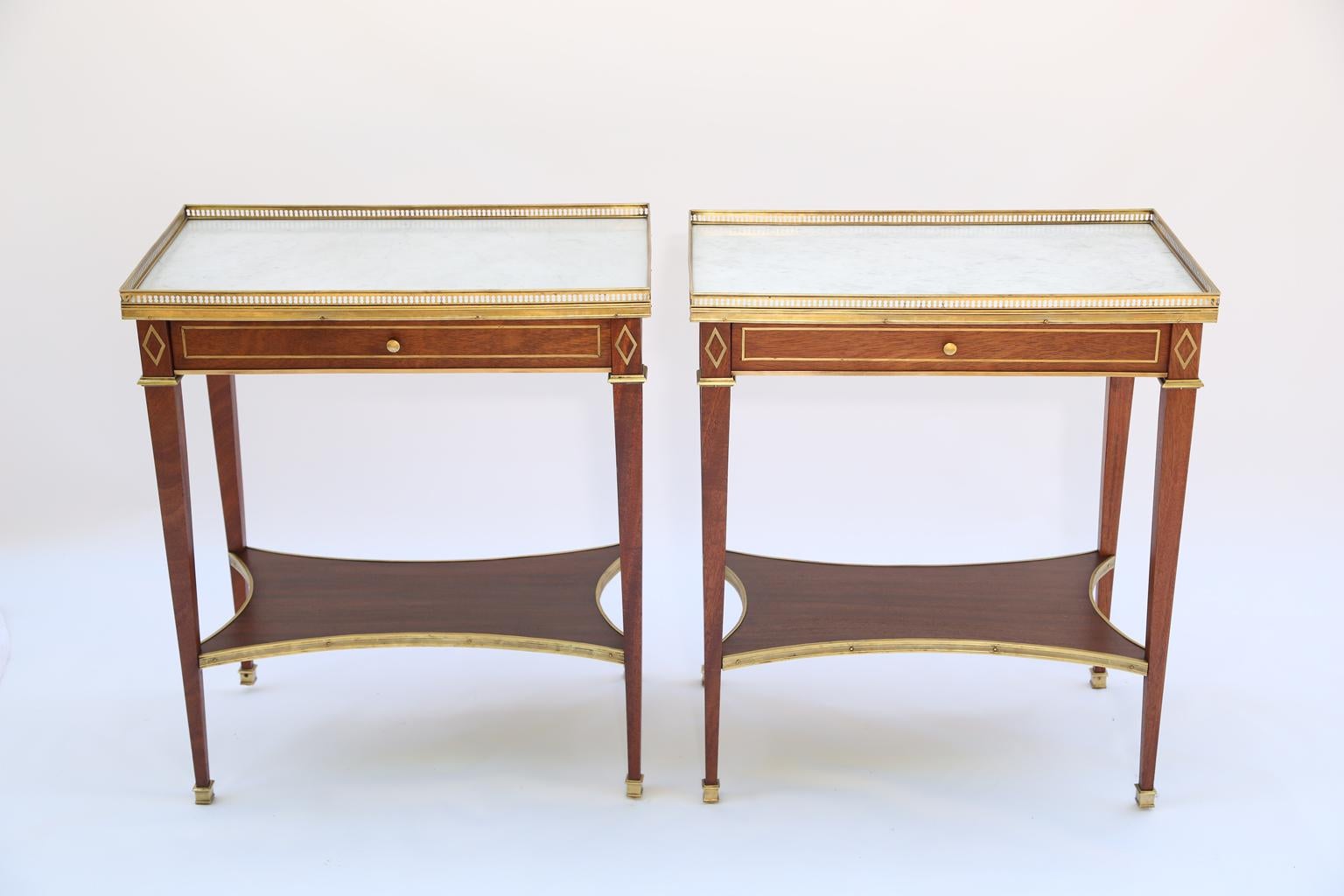 Pair of tables, in Directoire taste, of mahogany, each having rectangular tops of marble, surrounded by pierced gallery, single frieze drawer inset with gilt bronze stringing, and flanked by diamond corner accents, raised on square-section, tapering