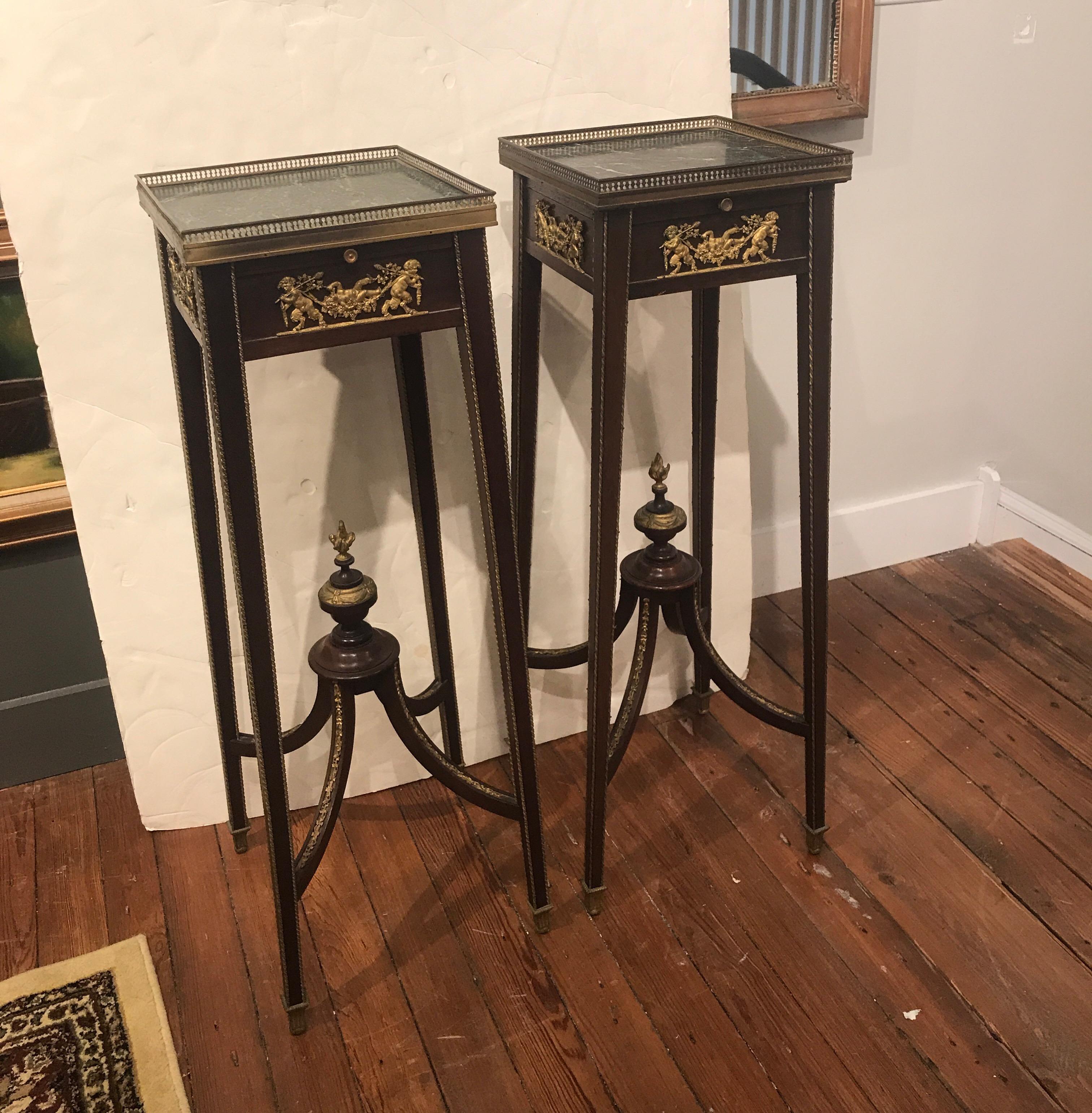 A pair of Directoire style mahogany marble and bronze pedestal stands. The green marble tops with aged gallery edge over four tapering splayed legs with elegant urn finial stretcher base. The top aprons with small pull out shelf in green leather,