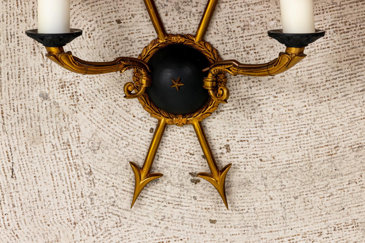 Circa 1960s pair of electrified painted brass sconces in the Directoire style that features a crossed arrow motif behind the double arm scrolled foliage shaped candle holder lights. The central ebonized dome is accented with a central star motif in