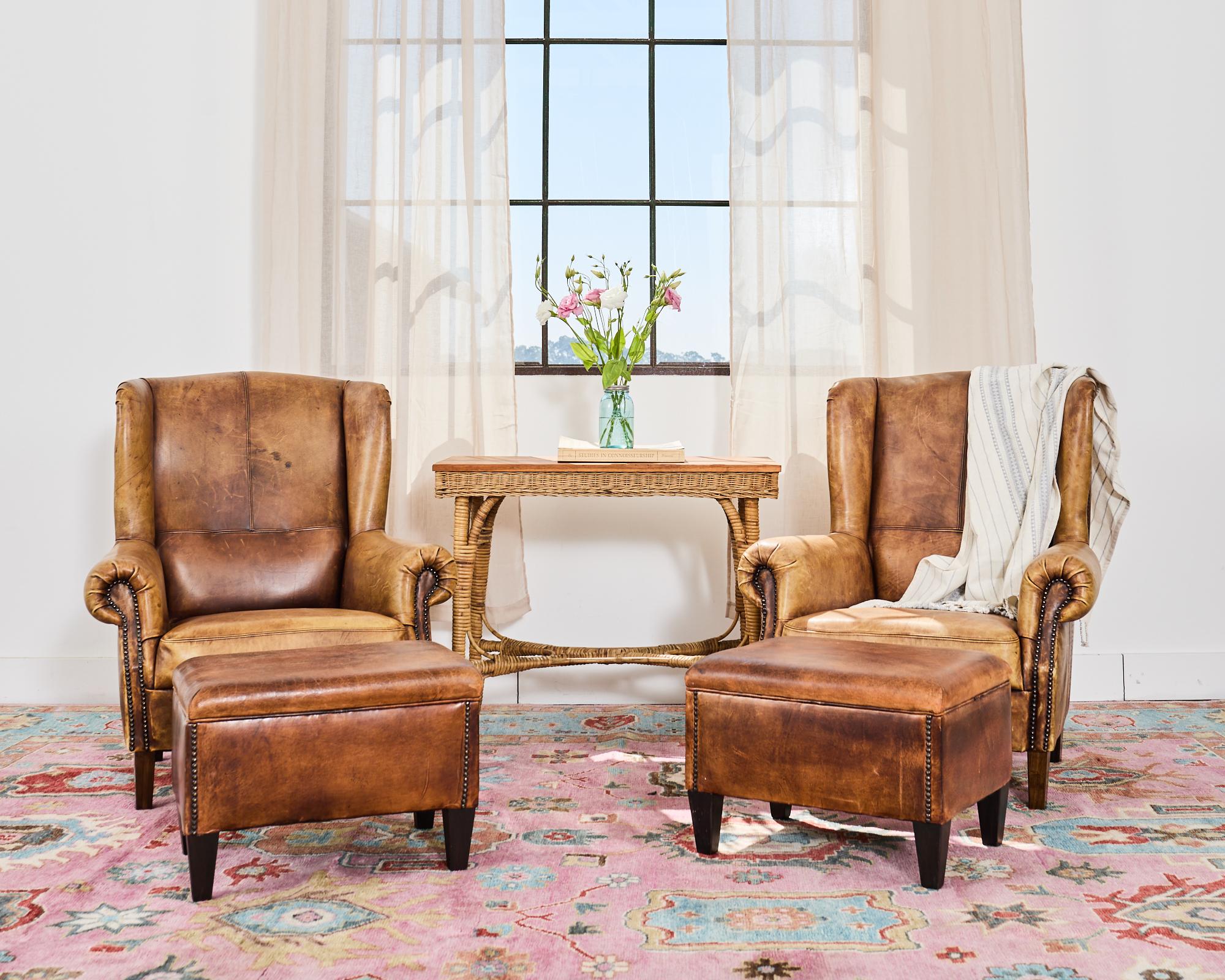 Handsome pair of rustic English style wingback lounge chairs or club chairs with ottomans. The chairs feature wood frames covered with aged, distressed cigar leather with patinated brass tack nail heads on the borders. The chairs have English style