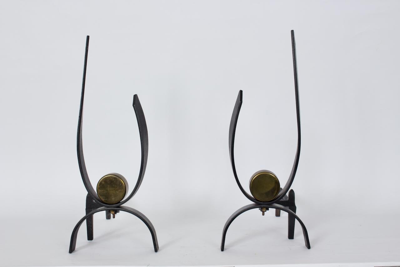 Modernist Donald Deskey Pair Brass & Black Cast Iron Fireplace Andirons.  Featuring a heavy balanced cast iron framework, u shape design in flat bar Iron strapping with patinated circular reflective Brass medallion details. The bolts to the rear