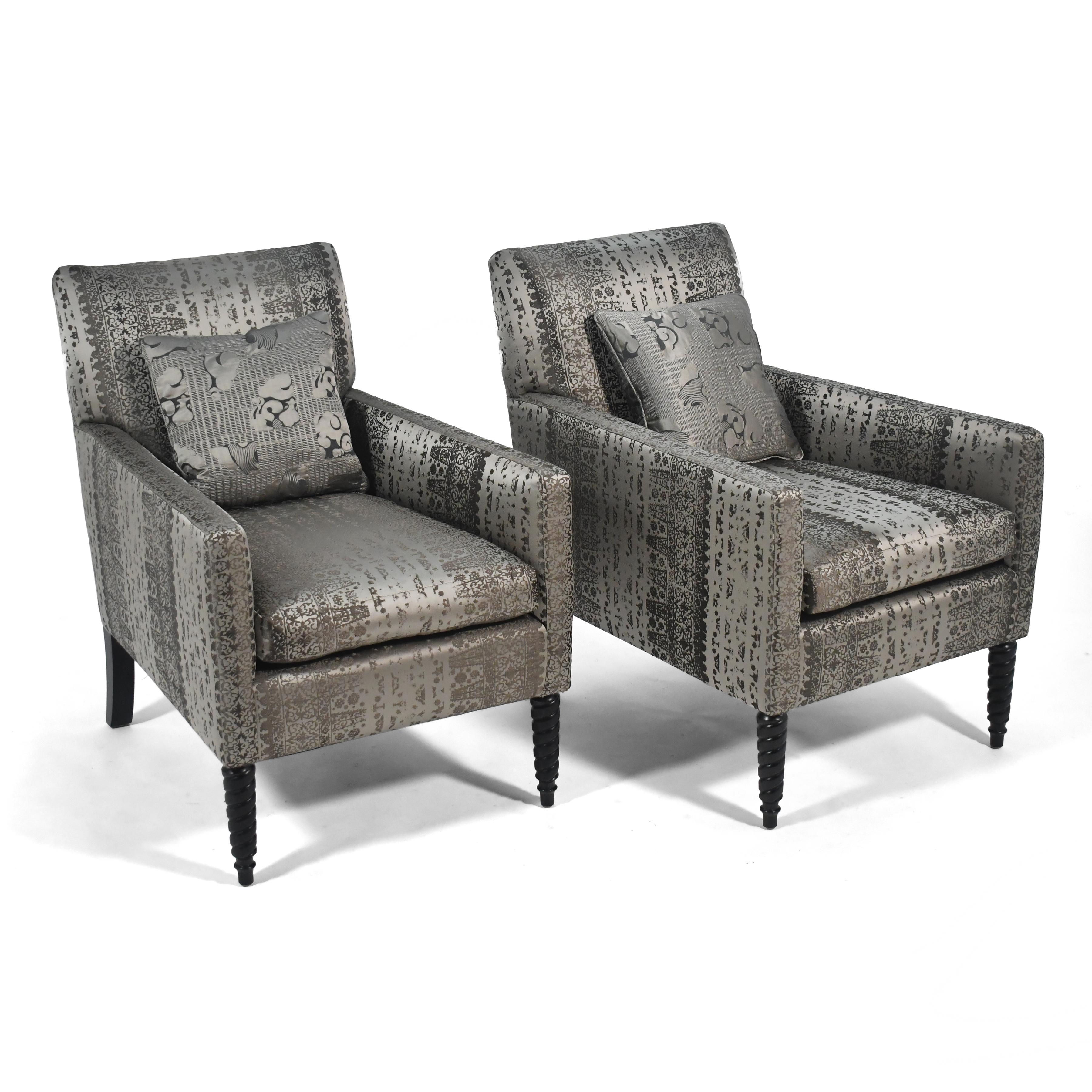 This pair of striking lounge chairs by Donghia feature crisply tailored upholstered bodies supported by sculptural legs– the front being turned with pleasing details, the rear with a sexy curve. The silver patterned upholstery adds an almost