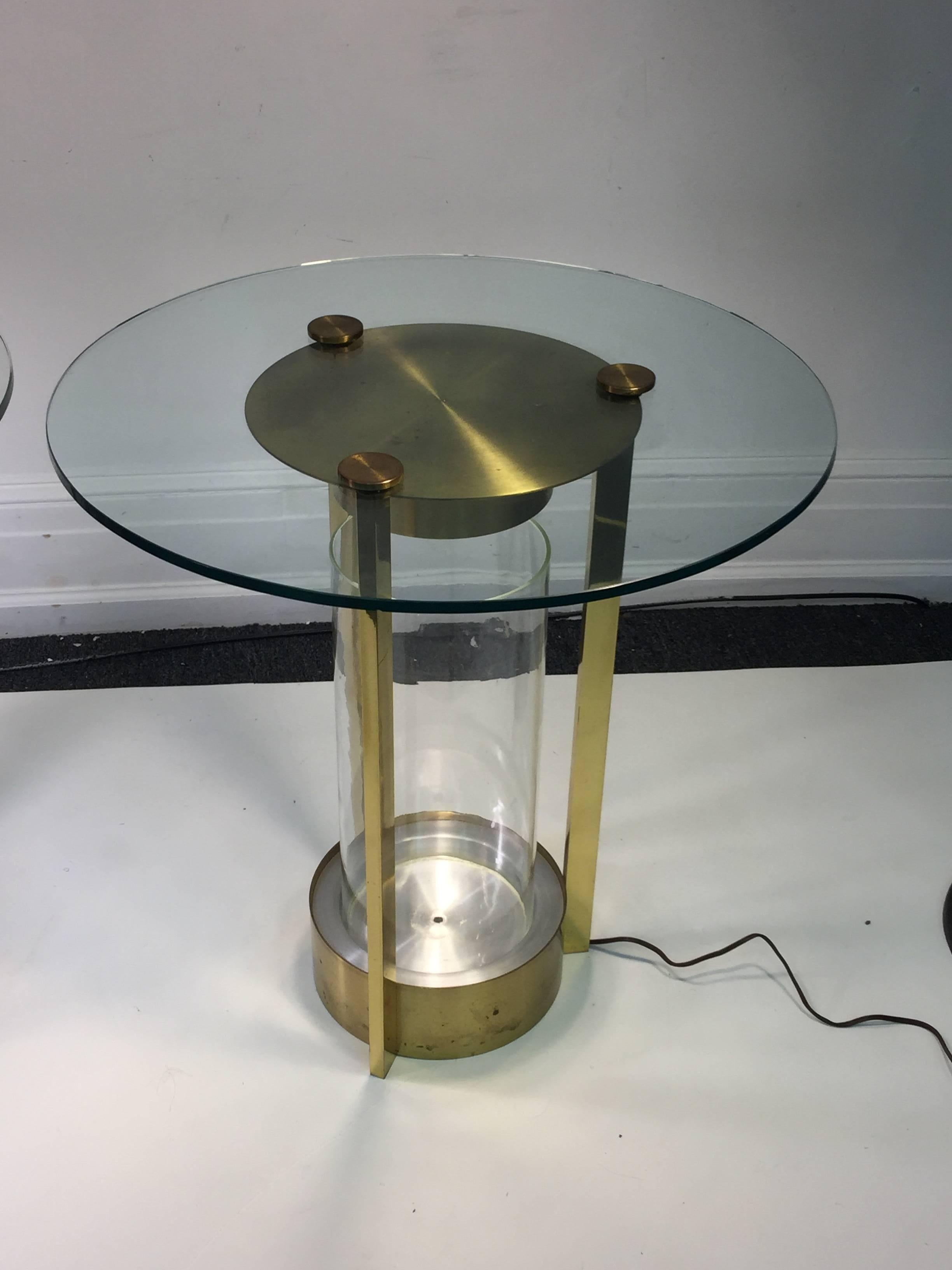 Great pair of modernist brass and glass end tables that illuminate when you would like with switch on the side under the round glass top. These are designed in the 1940s by designer Dorothy Thorpe. There is the original glass cylinder vase that fits