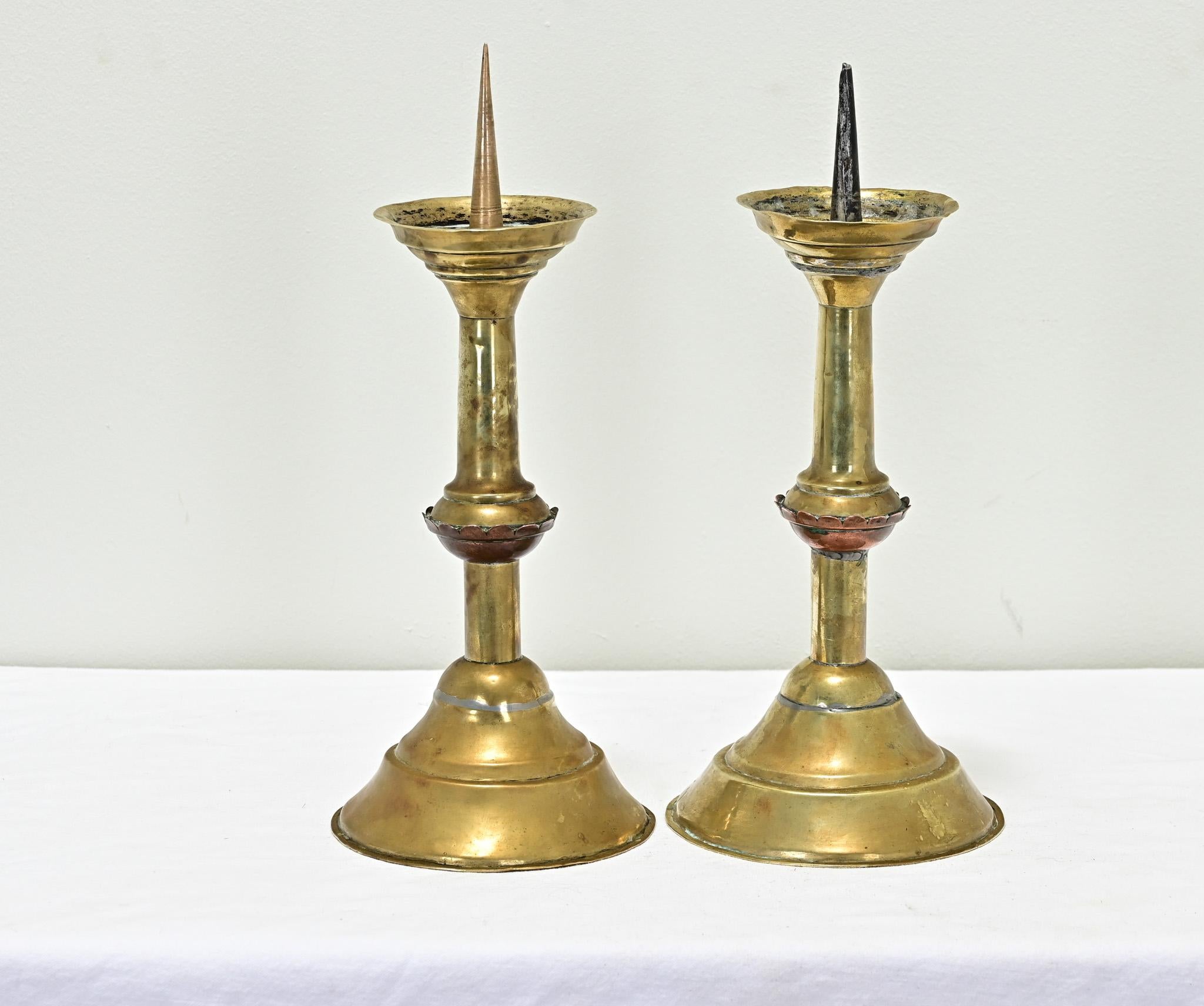 A pair of heavy brass candlesticks from the 1800’s have a rich, smooth patina. Primitive in design with large candle spikes for pillar wax candles. View all the detailed images to see the current condition.