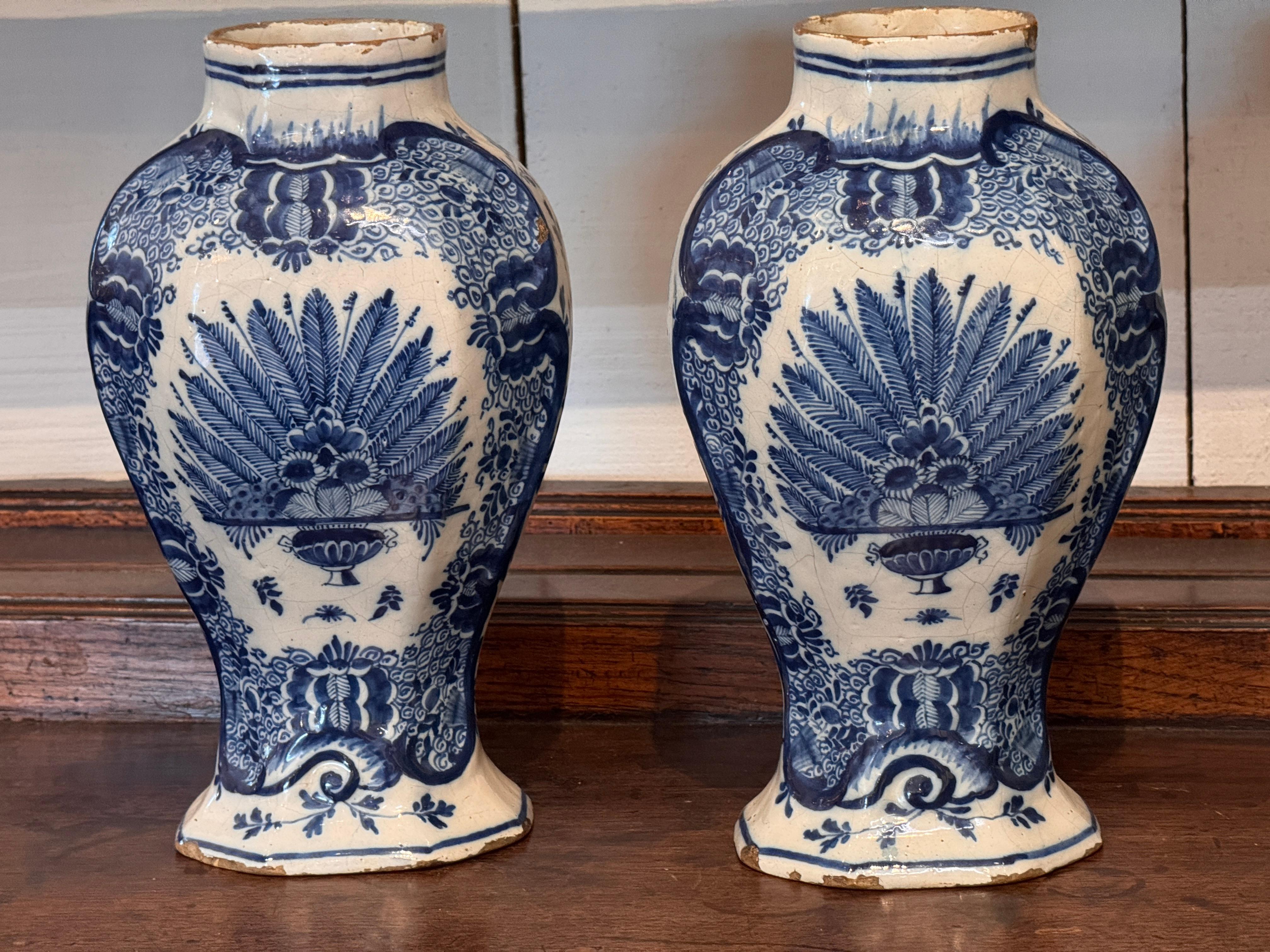 Paire de vases de Delft décorés en bleu et blanc de grandes gerbes de fleurs à plumes s'élevant d'urnes, encadrées par des bordures à motifs. La forme balustre repose sur une base évasée, avec une usure de la glaçure et des fritures le long des