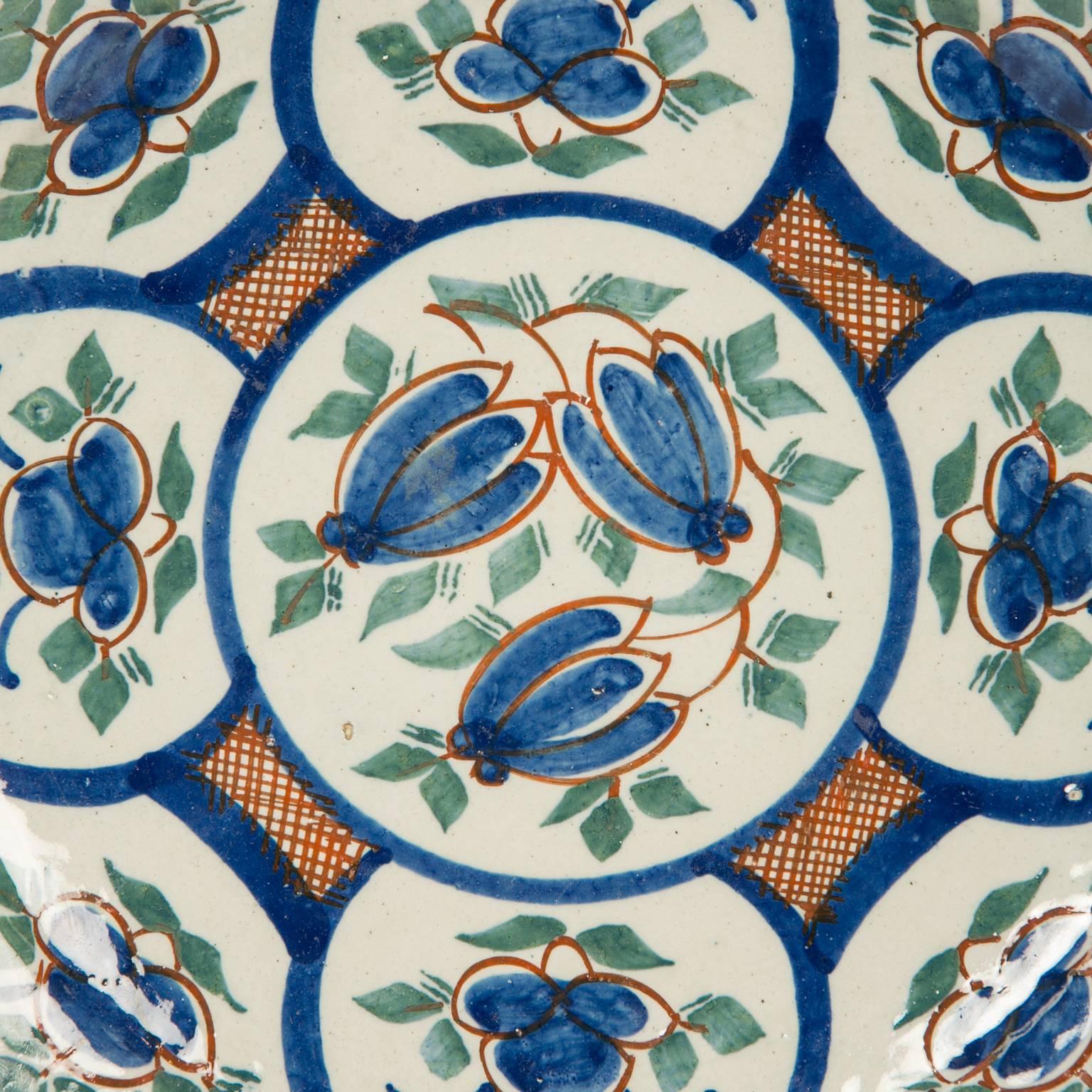 A pair of Dutch delft pancake plates decorated all-over with a colorful design of panels showing blue tulips and green leaves. The strong design is accented by areas of red cross hatching which complements the blue and green tulip decoration. 
These