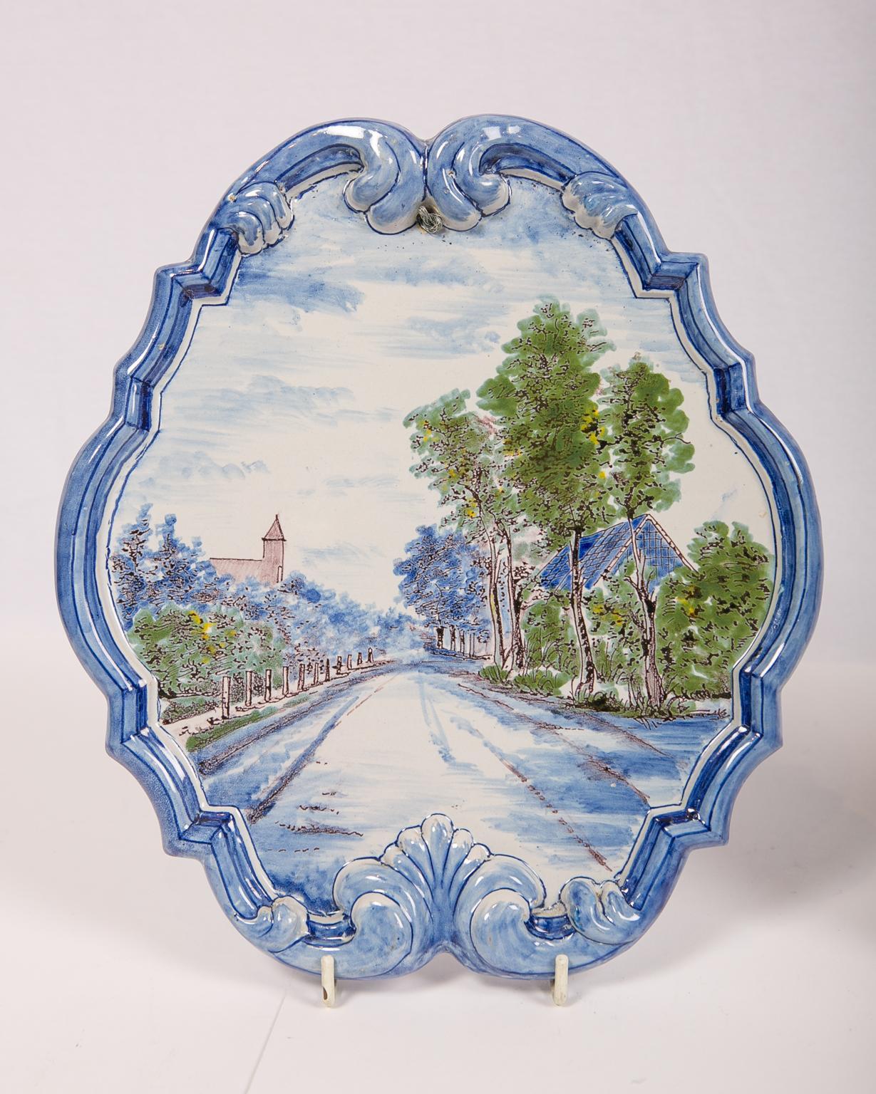A pair of Dutch delft wall plaques with blue raised borders each plaque showing a peaceful Dutch village scene; one shows a town from the waterfront with a towering church steeple and birds flying overhead: the other shows a long boulevard with