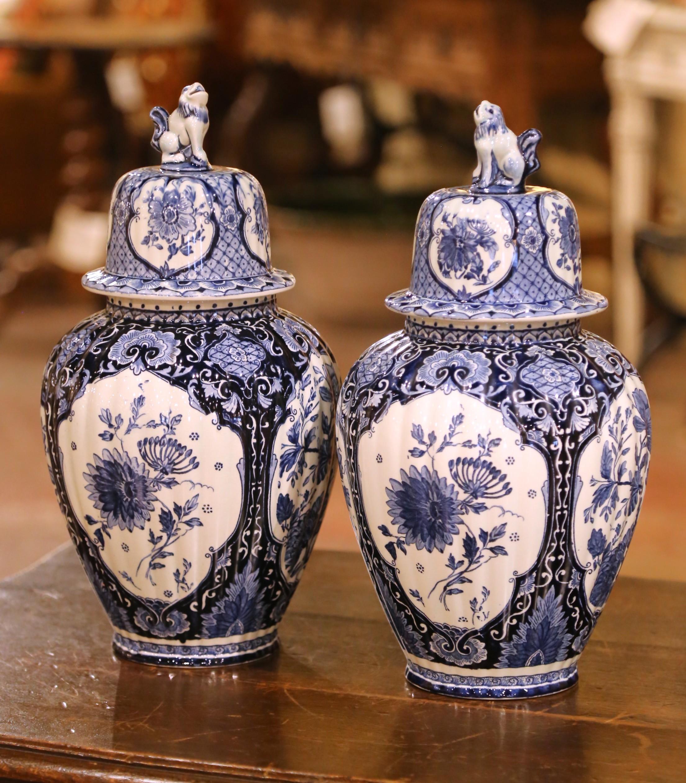 Enhance your collection with this exquisite pair of covered Delft vases. Crafted circa 1920 in Maastricht, Netherlands by Petrus Regout & Co. and-painted in the traditional blue and white palette, each faïence vase is adorned with scrolling floral