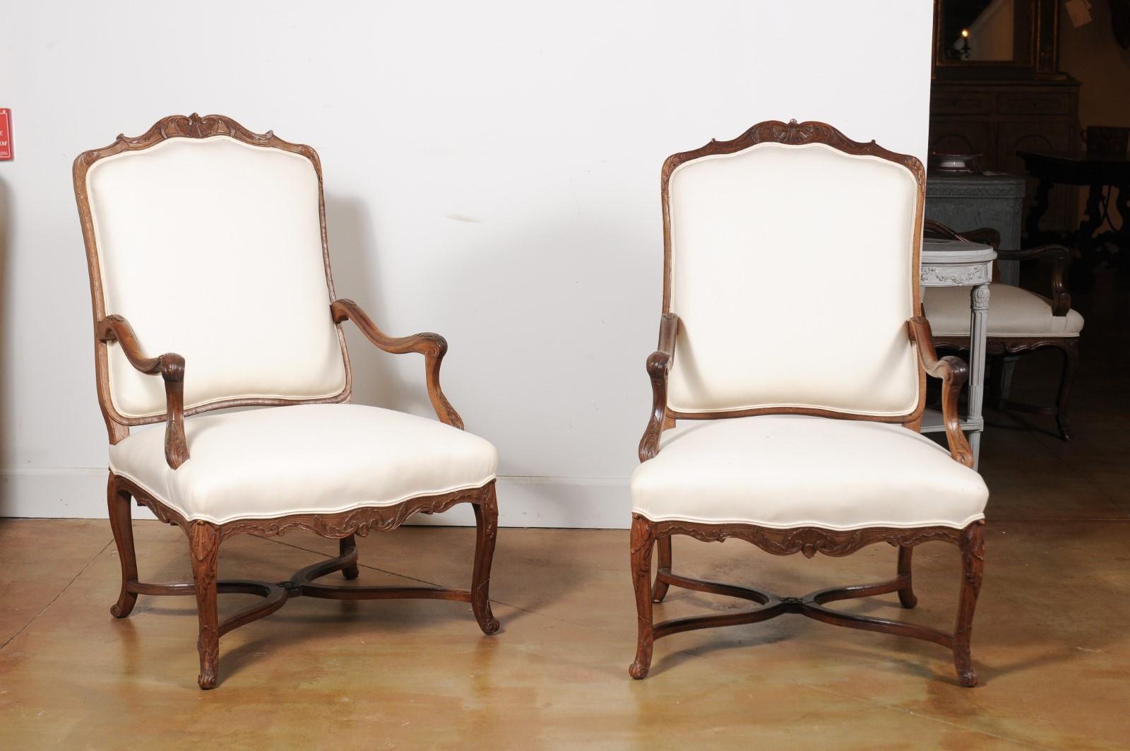 A pair of French Régence period walnut armchairs from the early 18th century, with carved crest, scrolling arms, cabriole legs and upholstery. Born in France during the transition of power between the Sun King and his heir Louis XV who was still too