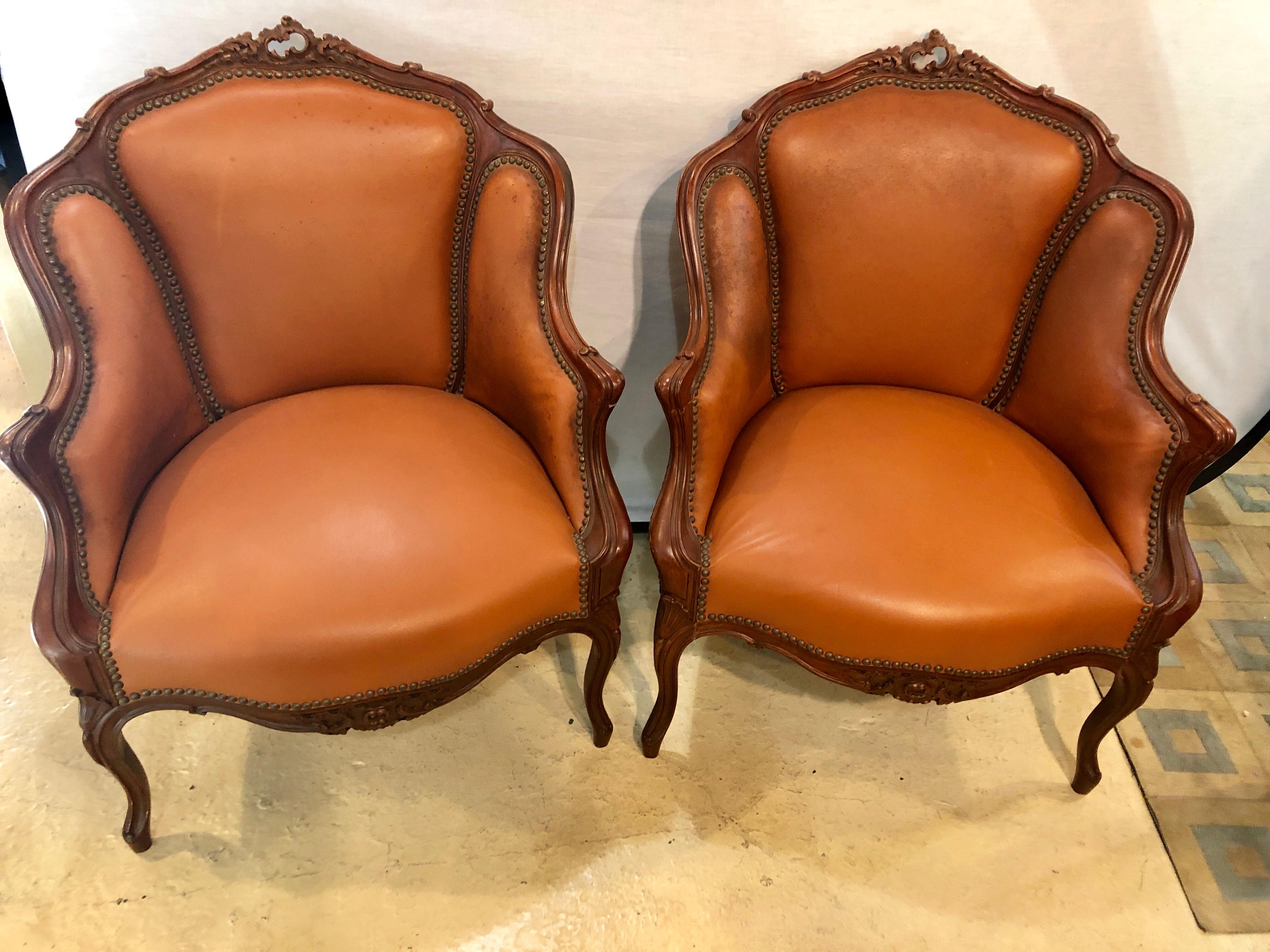 Pair of early 1920s Louis XV style barrel back bergere armchairs. Each in a worn tacked leather upholstery with stuffed sides, back and seats. The finely carved frames in a walnut finish with shell carvings. Upholstery is worn with light stains, no