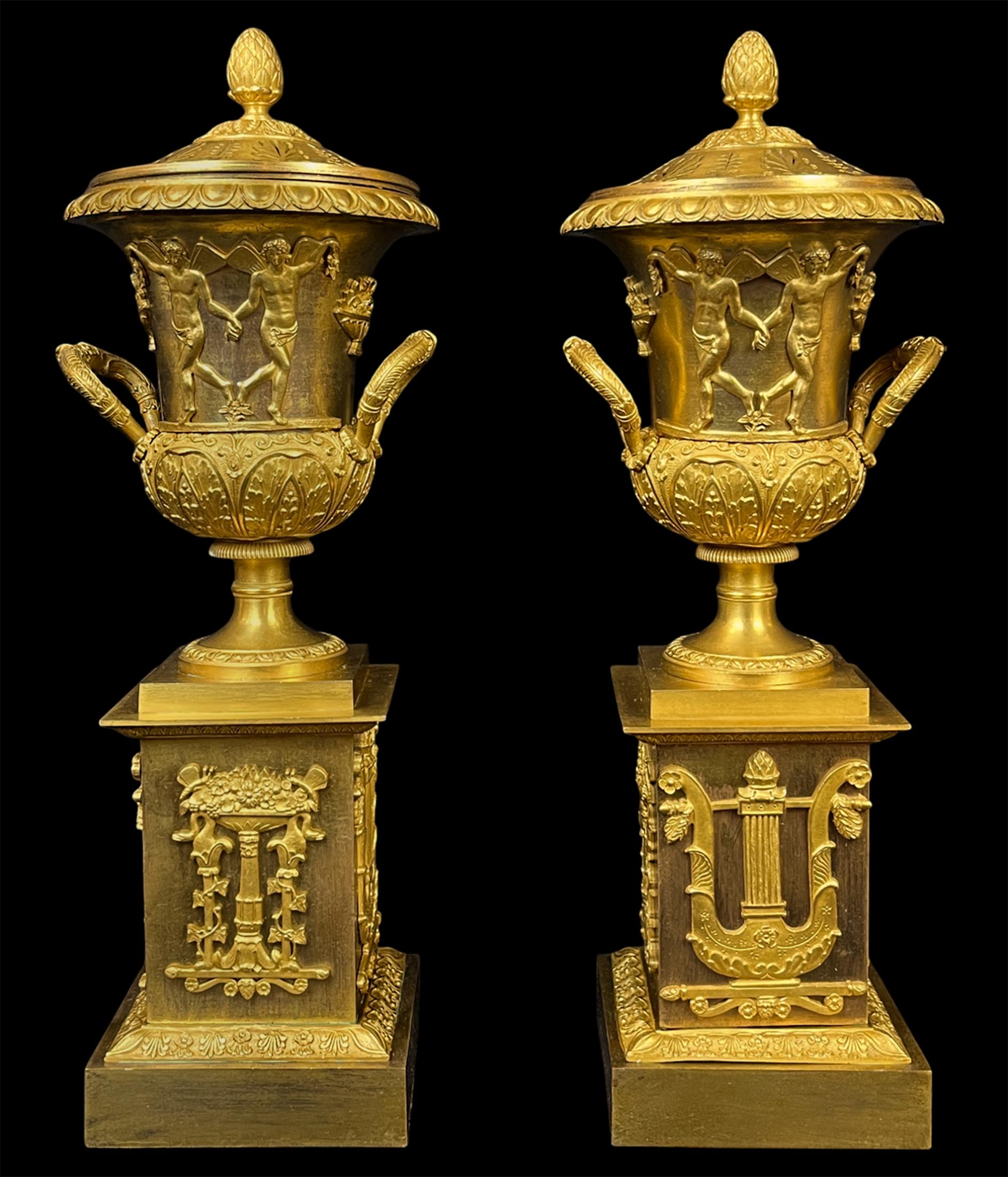 Pair of Early 19th C Empire Gilt Bronze Cassolettes. With a classic egg and dart pattern on the rim, the pair are adorned with two sets of winged male figures. The pair rest atop rectangular bases with lyres and jardinieres on the four faces.