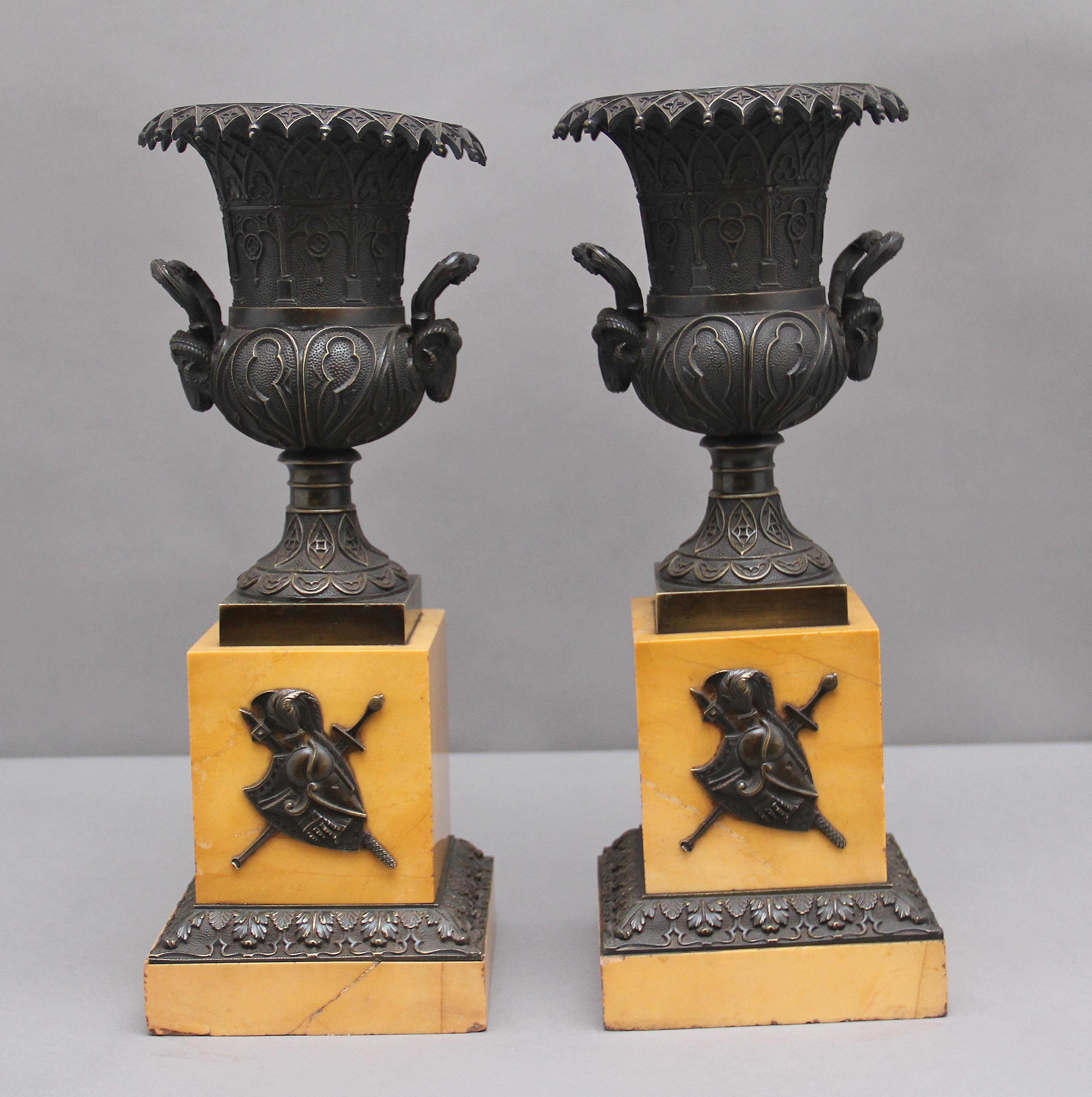 A pair of French early 19th century bronze urns on marble bases, the bronze engraved urns having wonderful intricate detail including rams head handles at either side of each urn, sitting on square column marble bases decorated with a coat of arms,
