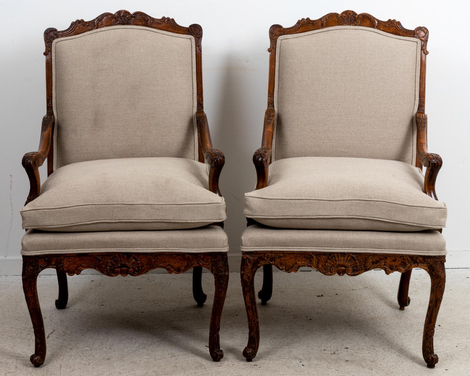 Circa 1800s pair of early 19th century French Louis XV style chairs on cabriole legs with carved top rail, bottom skirt, and scrolled arm holds ornamented with scrolled foliage and scallop shellwork. The chairs are freshly upholstered in linen with