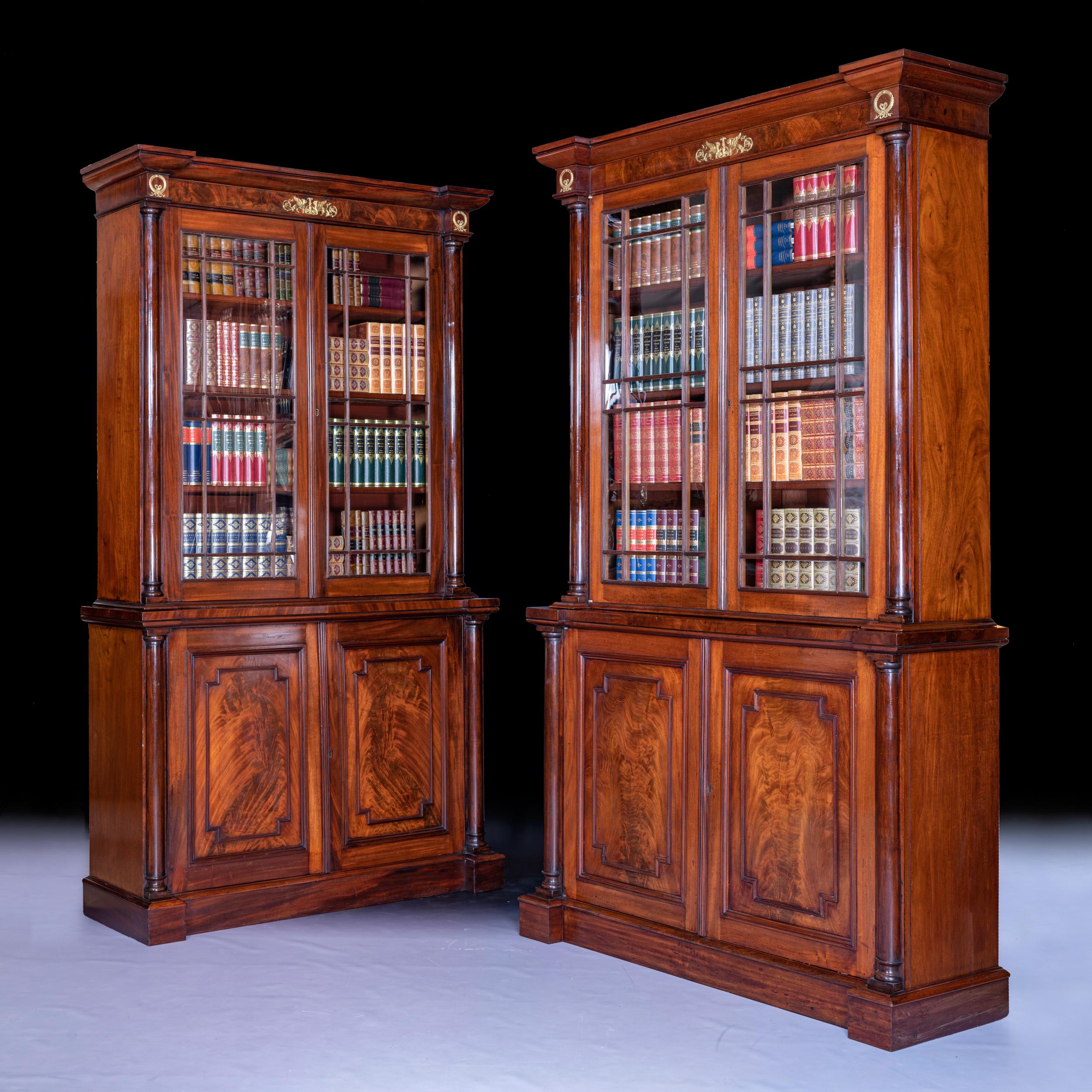 A pair of English Regency flame mahogany two door inverted breakfront bookcases attributed to Gillows of Lancaster, the top section with moulded cornice with applied ormolu plaques depicting opposing griffins above two geometric glazed doors with