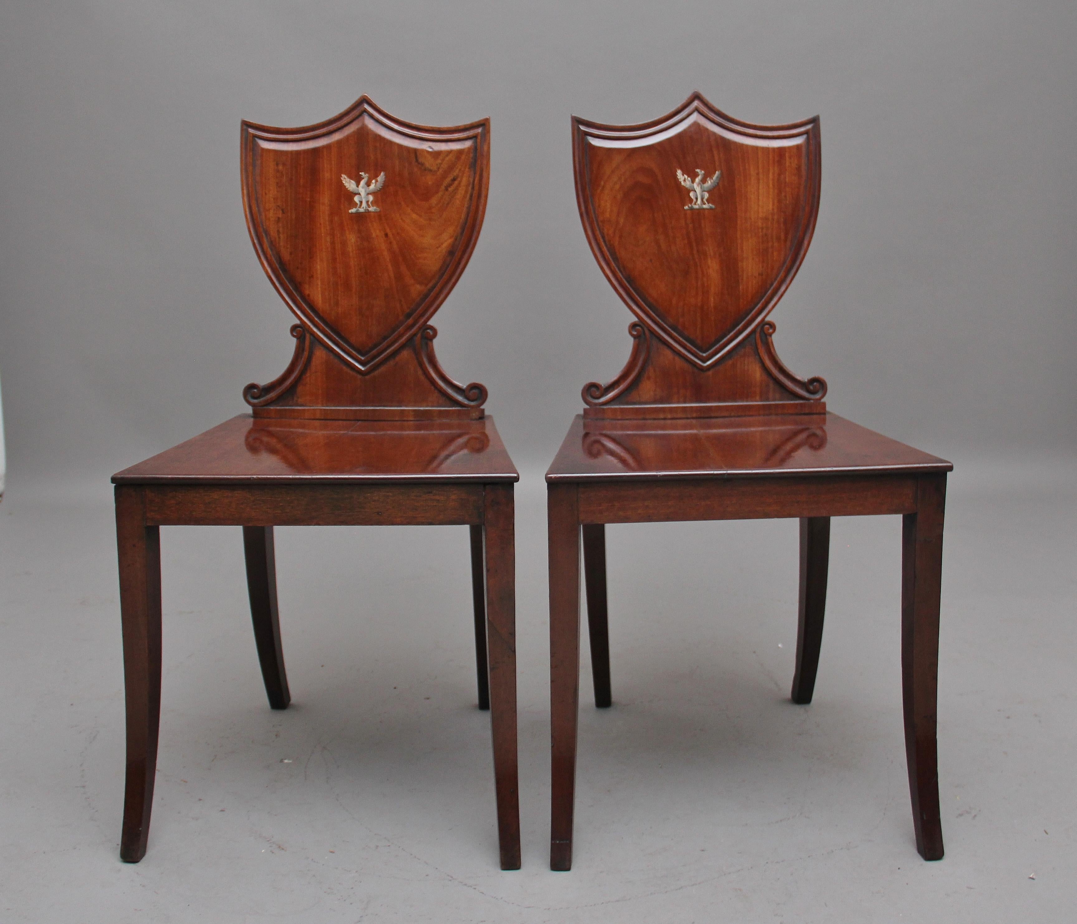 A pair of early 19th century mahogany hall chairs, having a shield back with a nice moulded edge, with a griffin emblem at the centre of the shield, leading down to a shaped scroll support onto the hard wood seat, supported on swept rear and front