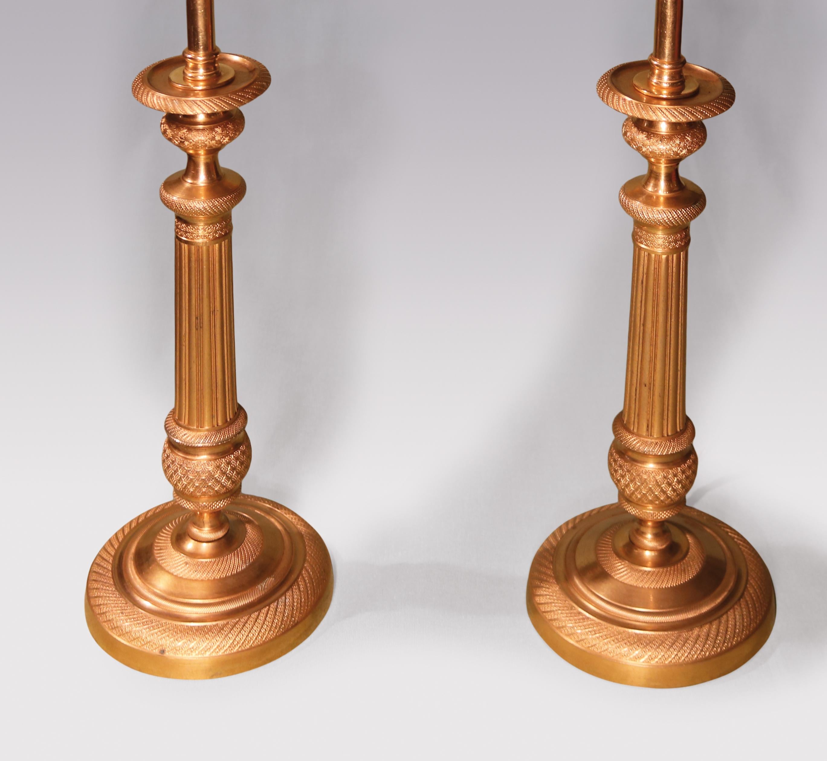 A pair of early 19th century ormolu candlesticks engine-turned throughout having urn shaped nozzles above reeded stems with circular bases. (Now converted to lamps) Height with shades: 20.75'.