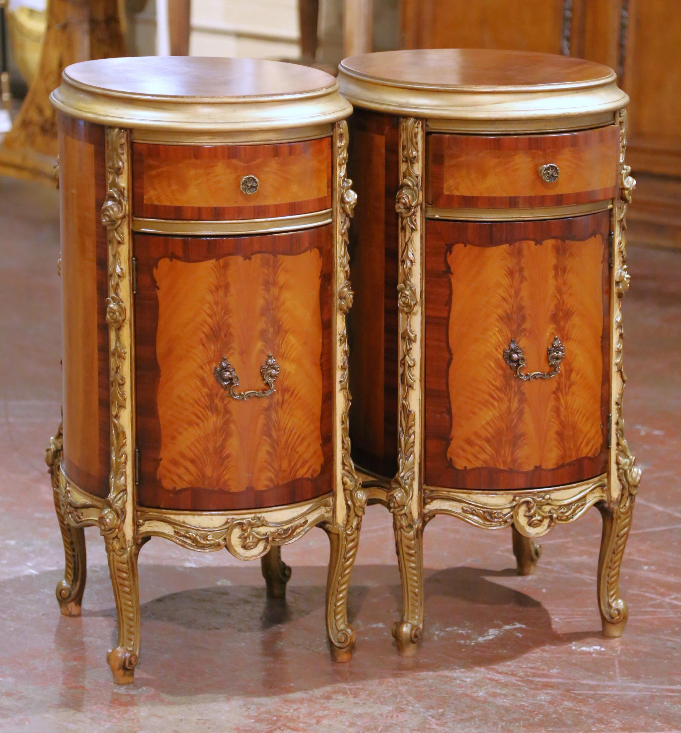 Crafted in France circa 1930, this refined pair of Louis XV style oval cabinets showcases richly figured flame mahogany accented with delicate marquetry. Each cabinet features a bookmatched veneer oval top framed with a molded edge above a single