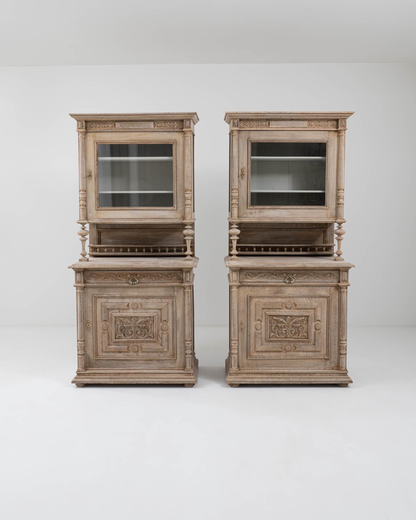 These pair of 1900s oak Belgian vitrines à deux corps flaunt the eclectic richness of their decorative elements. These display cabinets boast neoclassical shaped cornices, embossed foliate carvings, and masterfully crafted balusters; visually