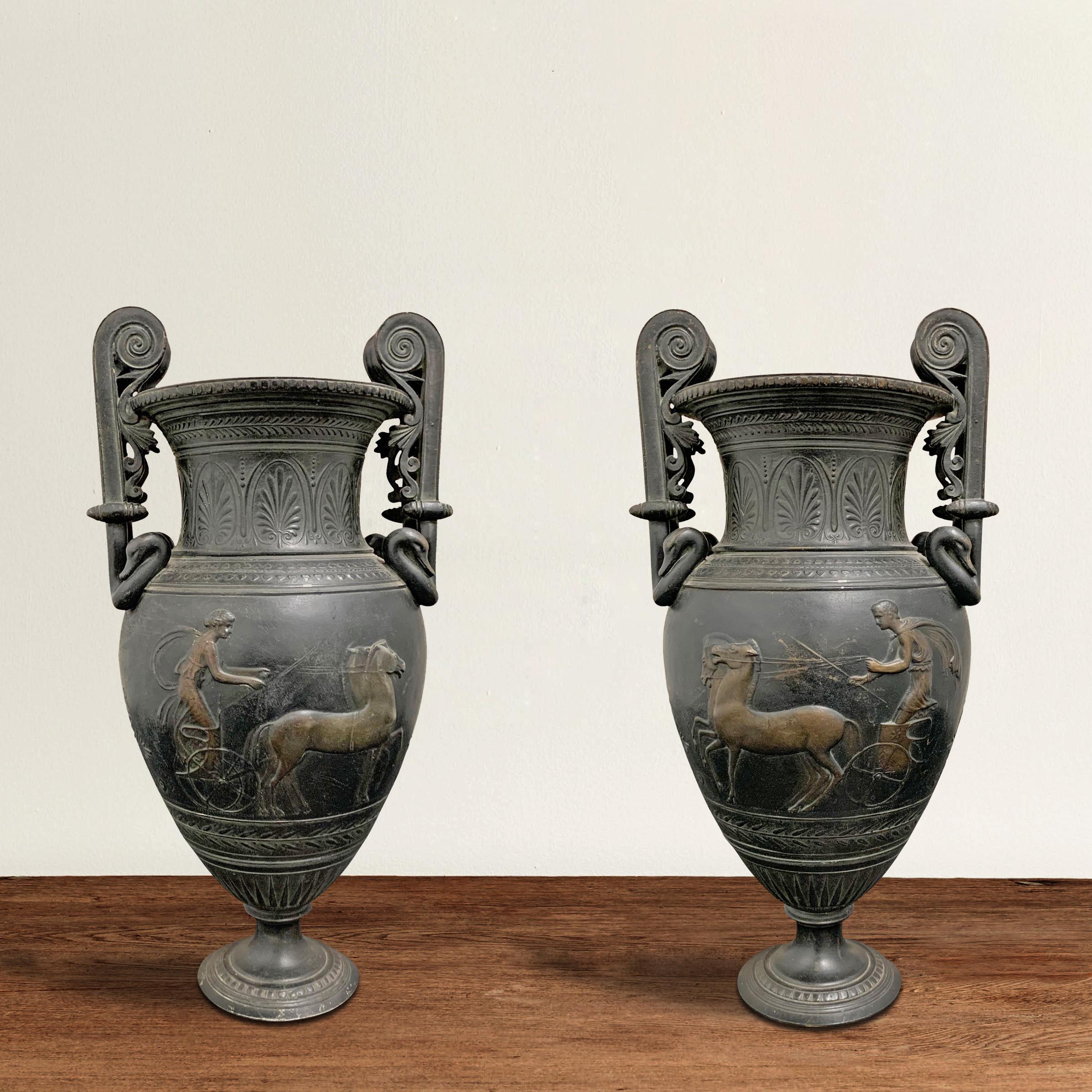 A phenomenal pair of early 20th century French cast bronze Roman-style urns with volute handles decorated with goose heads, and a frieze in low-relief depicting a man and woman chasing each other in chariots being pulled by horses. Classical floral