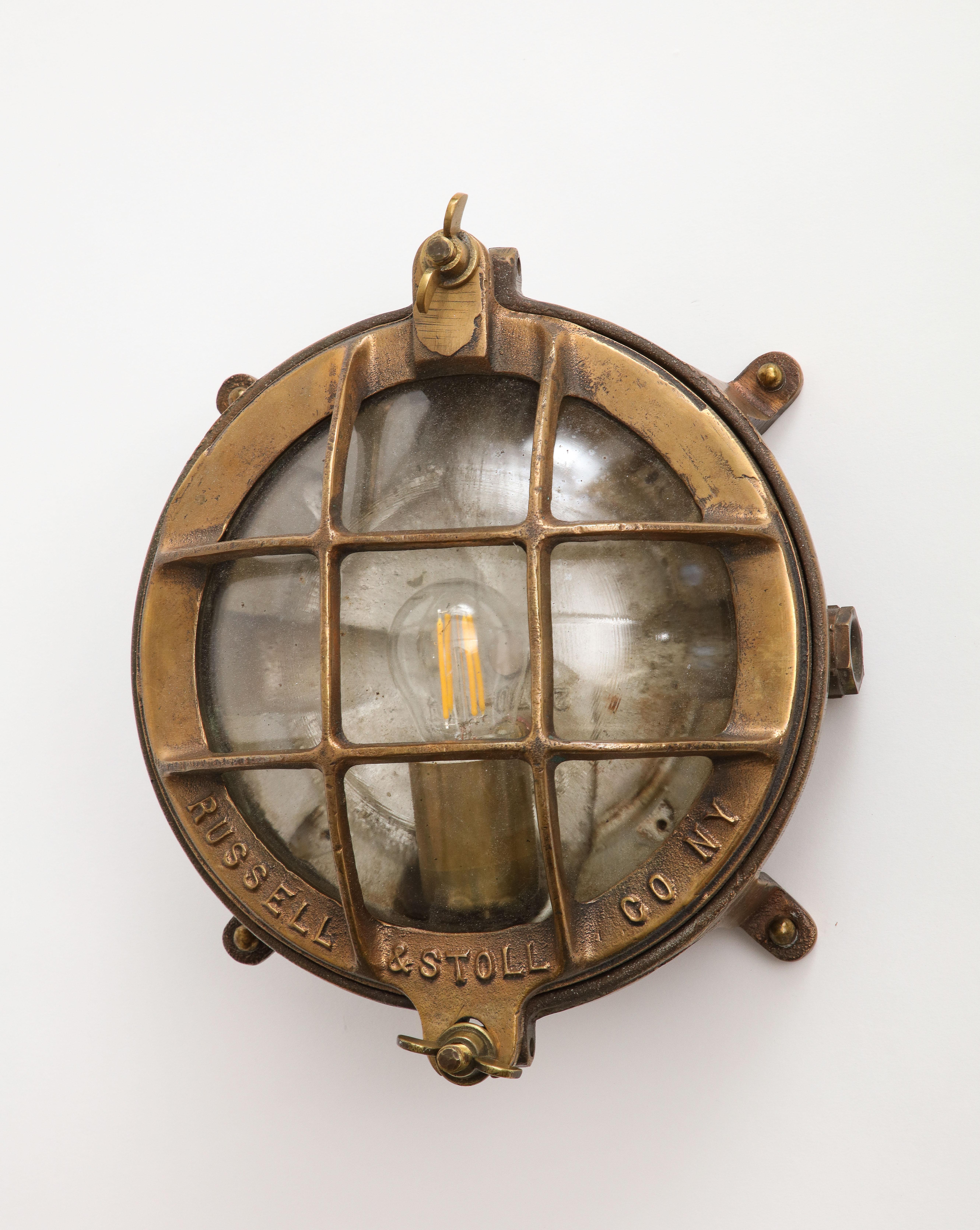 A pair of early 20th century Russell & Stoll Co. cast bronze lamps, can be wall mounted as a sconce or flush mount on the ceiling. The lamps are on a footed base with the glass dome protected by a well designed bronze cage. Industrial period, ship