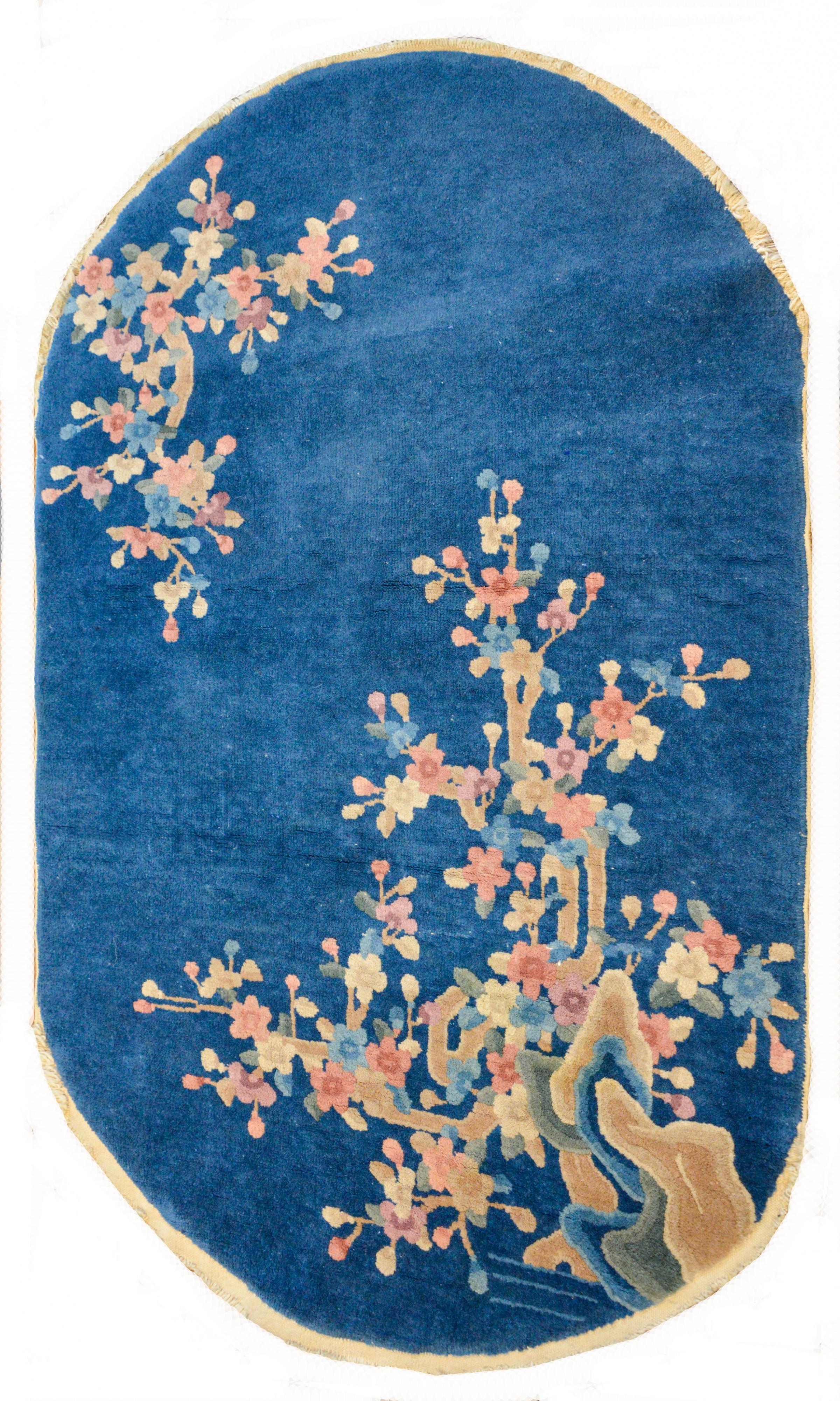 A pair of early 20th century Chinese Art Deco oval rugs, each with a flowering cherry blossom tree growing from within a scholar's rock in each corner, and set against a light indigo background.