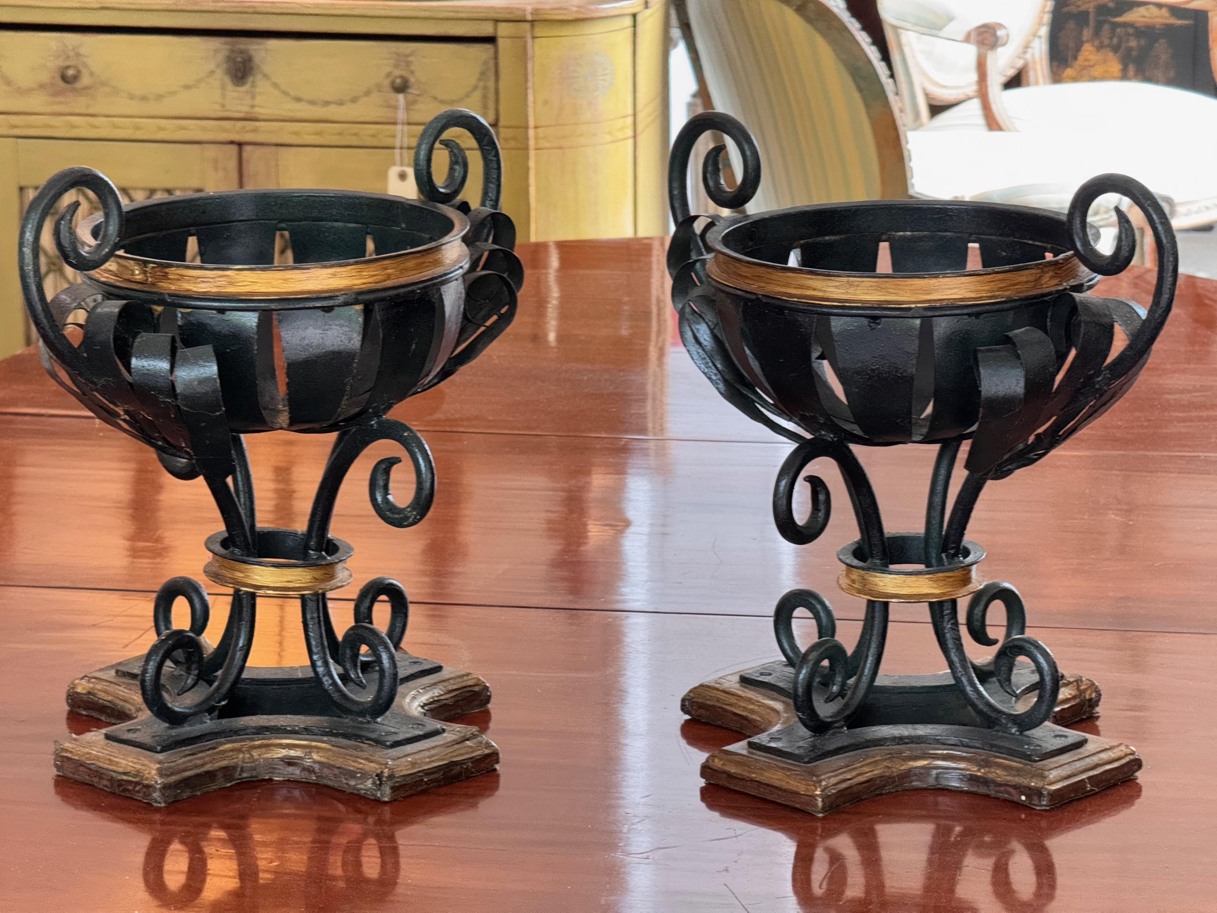 Pair of sculptural metal urns with open slat design and ornate scrolled handles. Finished in black with gilded accents and mounted on shaped wooden bases. Bold and architectural, these urns add classical form and contrast to interior or covered