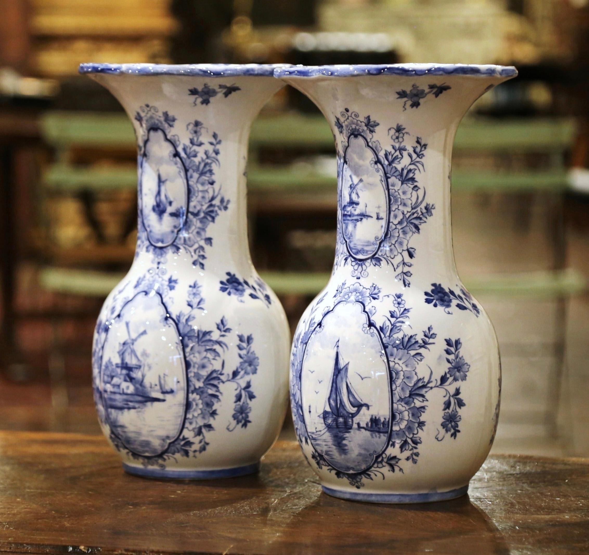 Decorate a shelf or console table with this elegant pair of antique Delft vases. Crafted in Holland circa 1920, both tall vases with wide mouth, feature a scalloped rim at the top over a long neck and round shape bottom. Each vessel is hand painted