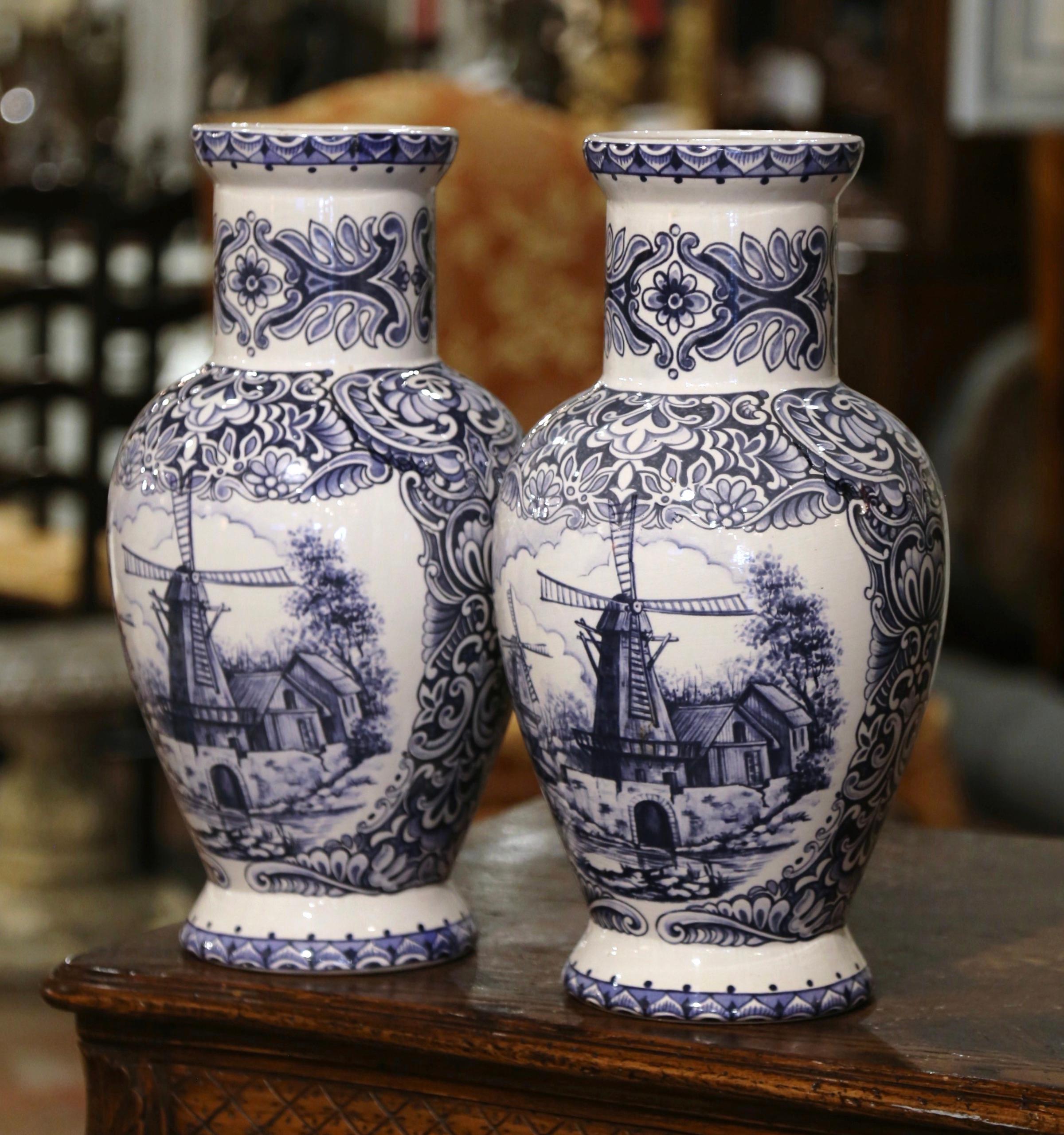 Decorate a shelf or console table with this elegant pair of antique Delft vases. Crafted in Holland circa 1920, both urns are round in shape and feature a long neck ending with a wide mouth at the pediment. Each vessel is hand painted in the