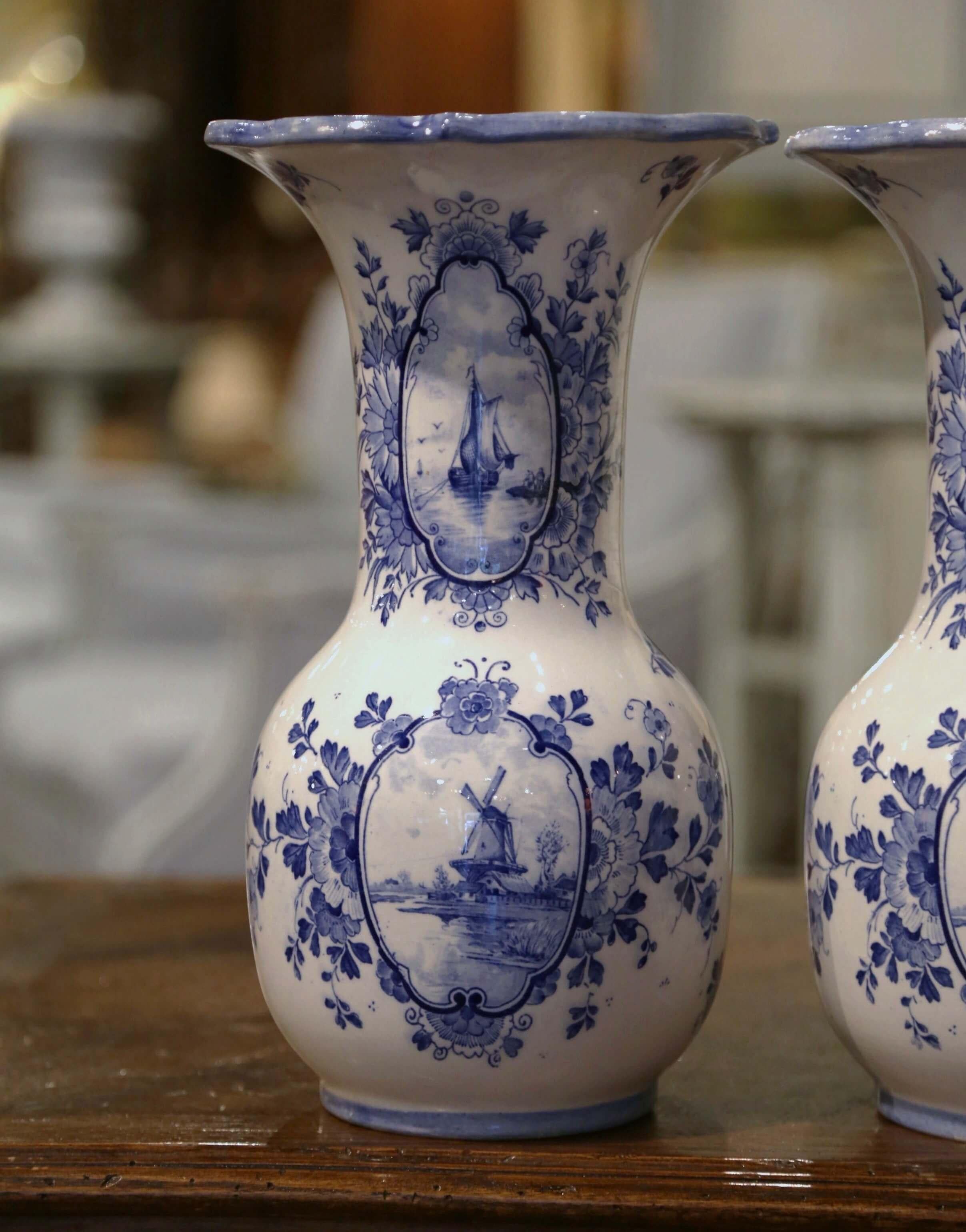 Decorate a shelf or console table with this elegant pair of antique Delft vases. Crafted in Holland circa 1920, both vases with wide mouth feature a scalloped rim at the top over a long neck and round shape bottom. Each vessel is hand painted in the