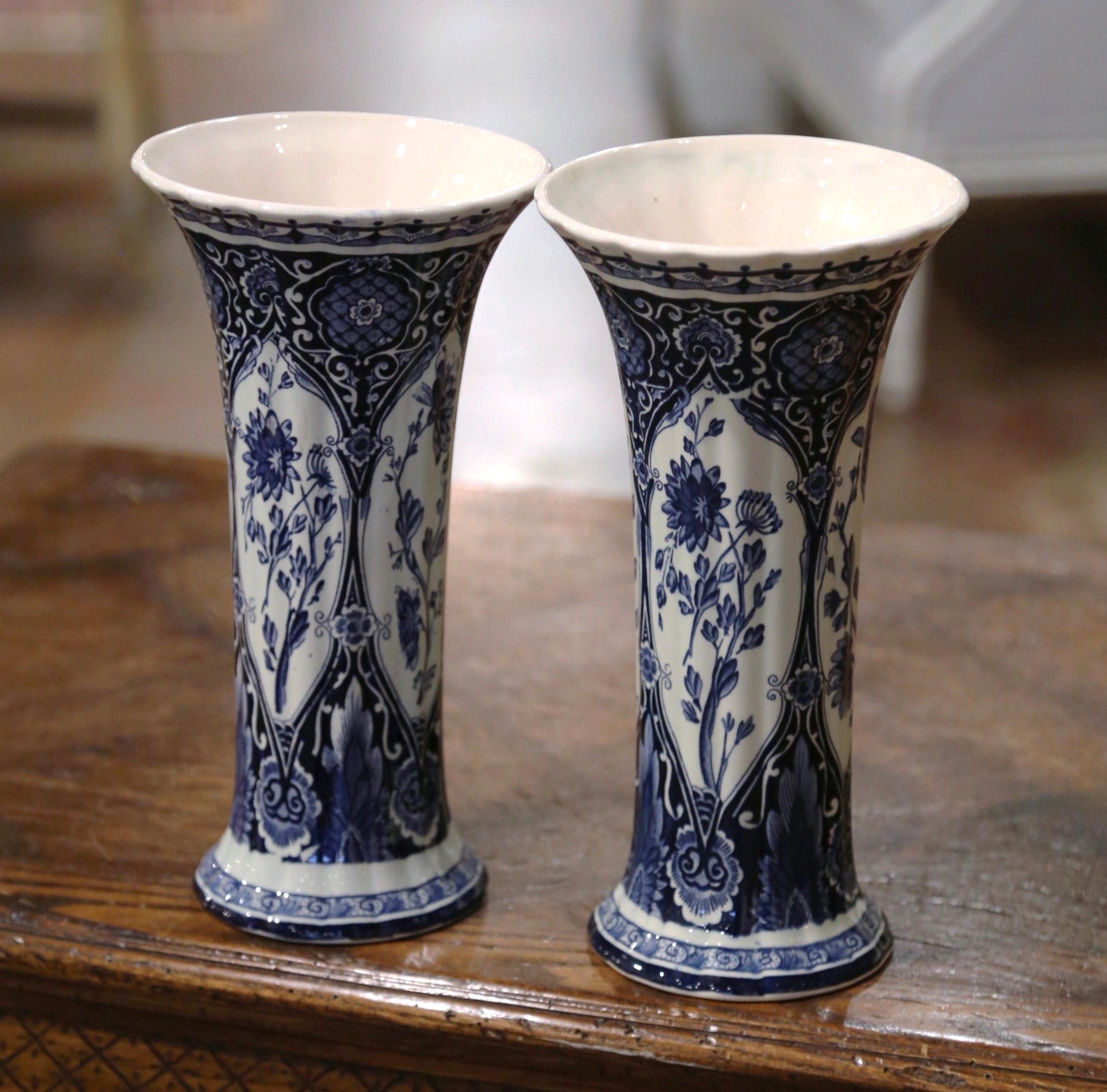 Decorate a shelf or console table with this elegant pair of antique Delft style vases. Crafted in Netherlands circa 1920, the hand-painted trumpet vases are round in shape over a long, elegant, fluted neck. Each vintage vessel is painted in the blue