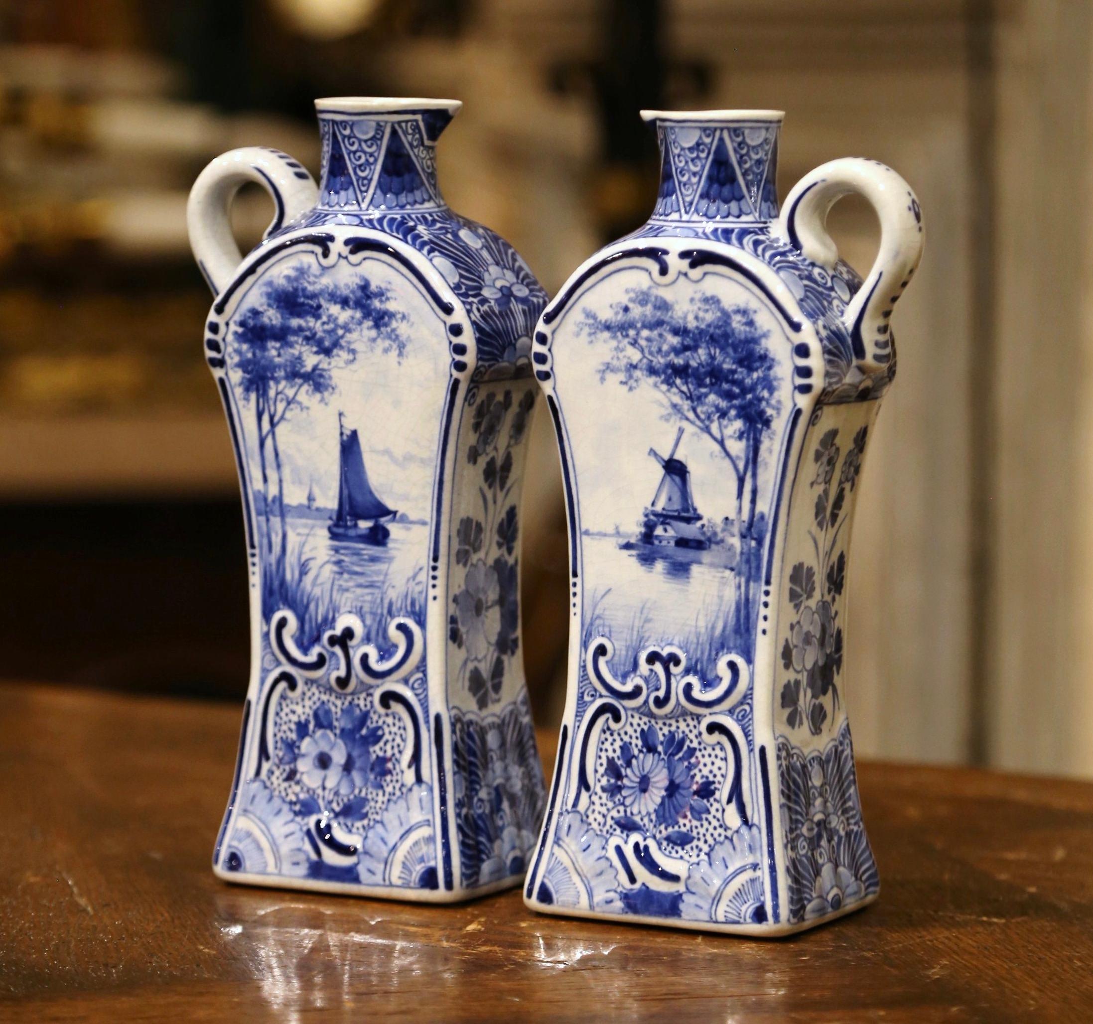 Decorate a shelf or kitchen counter with this elegant pair of antique Delft olive oil or vinegar containers. Crafted in Holland circa 1920, both tall vases feature an intricate shape and are dressed with a side handle. Each vessel is hand painted in