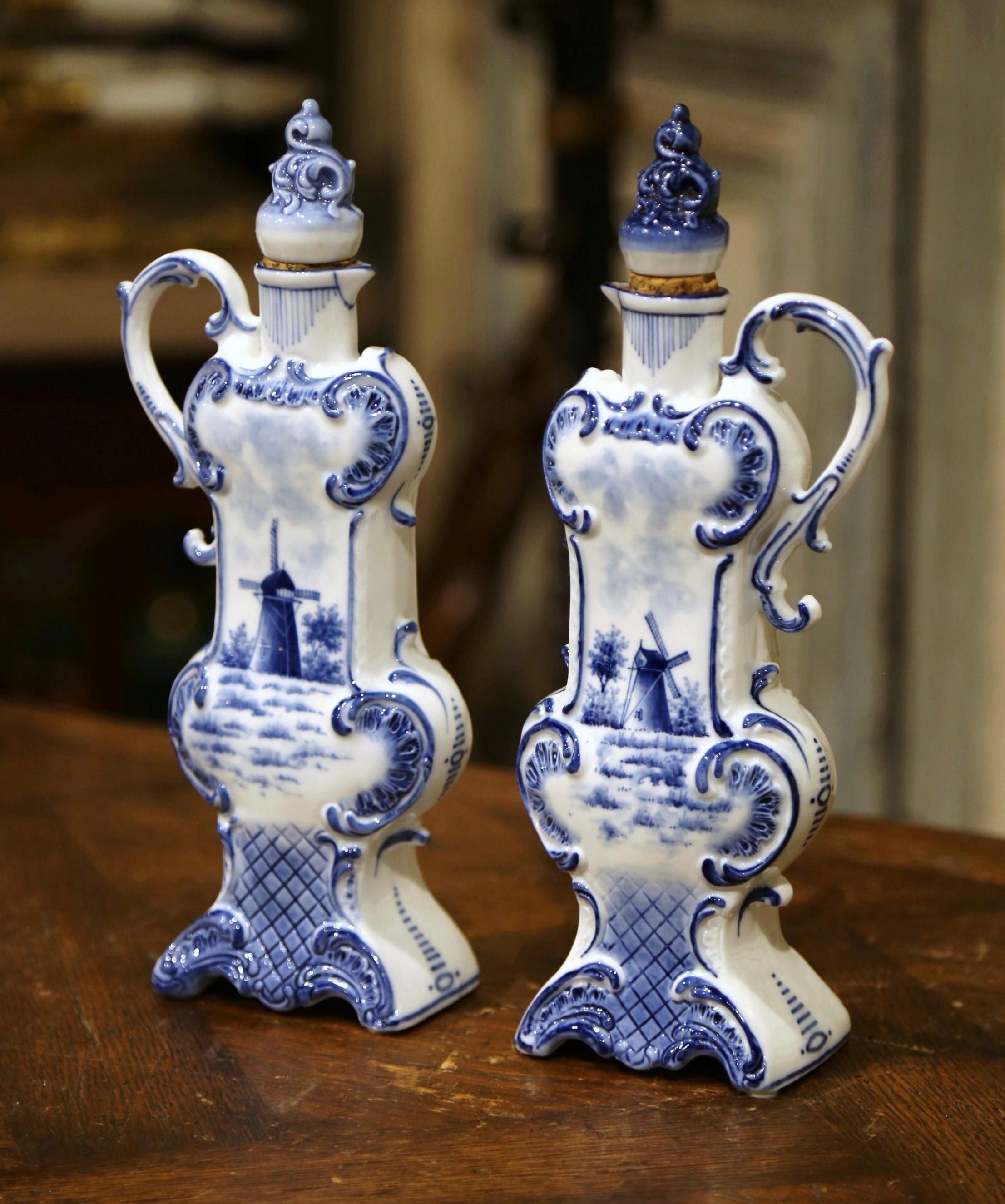 Decorate a shelf or kitchen counter with this elegant pair of antique Delft olive oil containers. Crafted in Holland circa 1920, both tall vases with cork tops, feature an intricate shape and are dressed with a side handle. Each vessel is hand