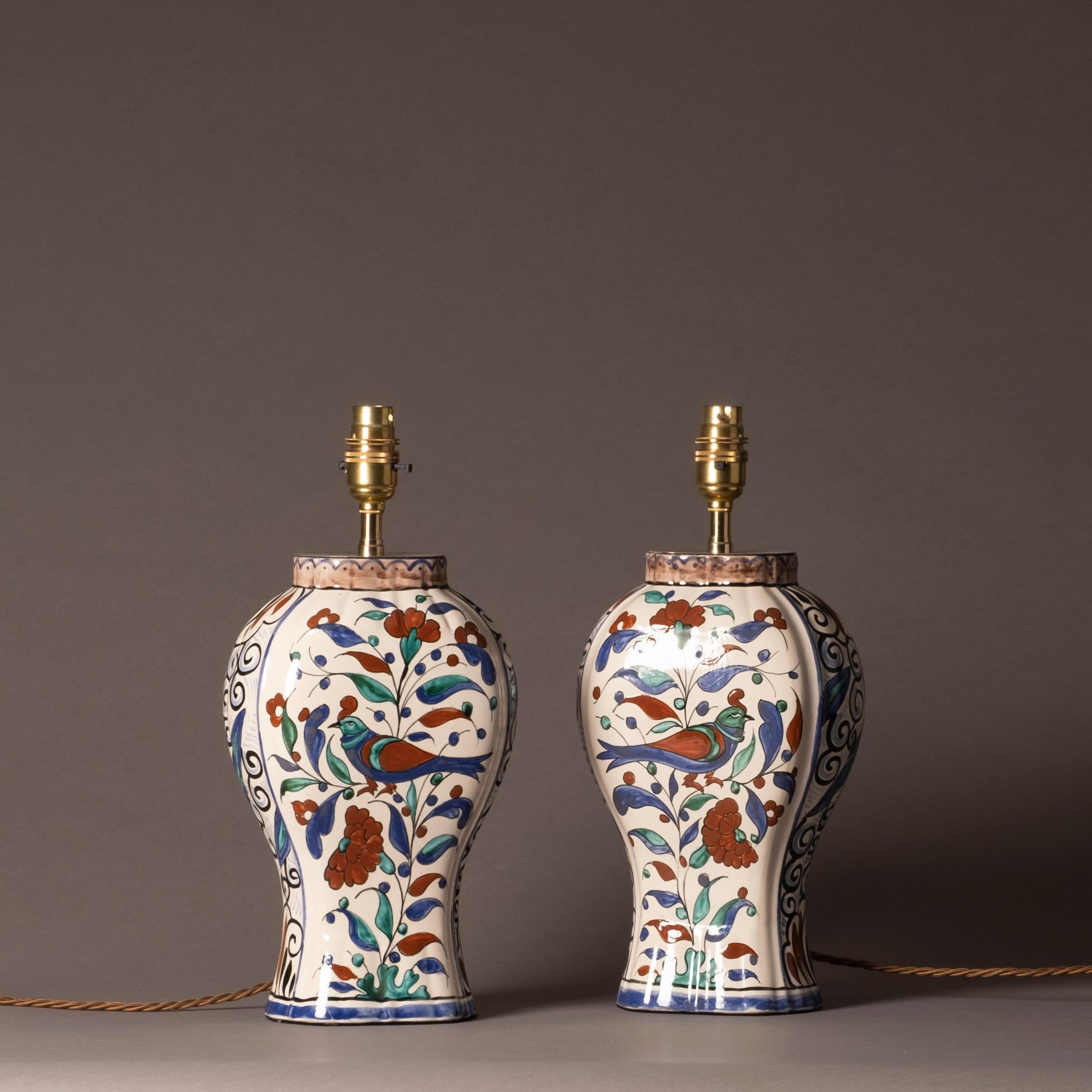 A pair of early 20th century faience pottery vases, of baluster form, the bodies decorated with birds, foliage and scrollwork in red, green, blue and black glazes upon a white ground. Now mounted as a pair of table lamps.

Height dimensions refer