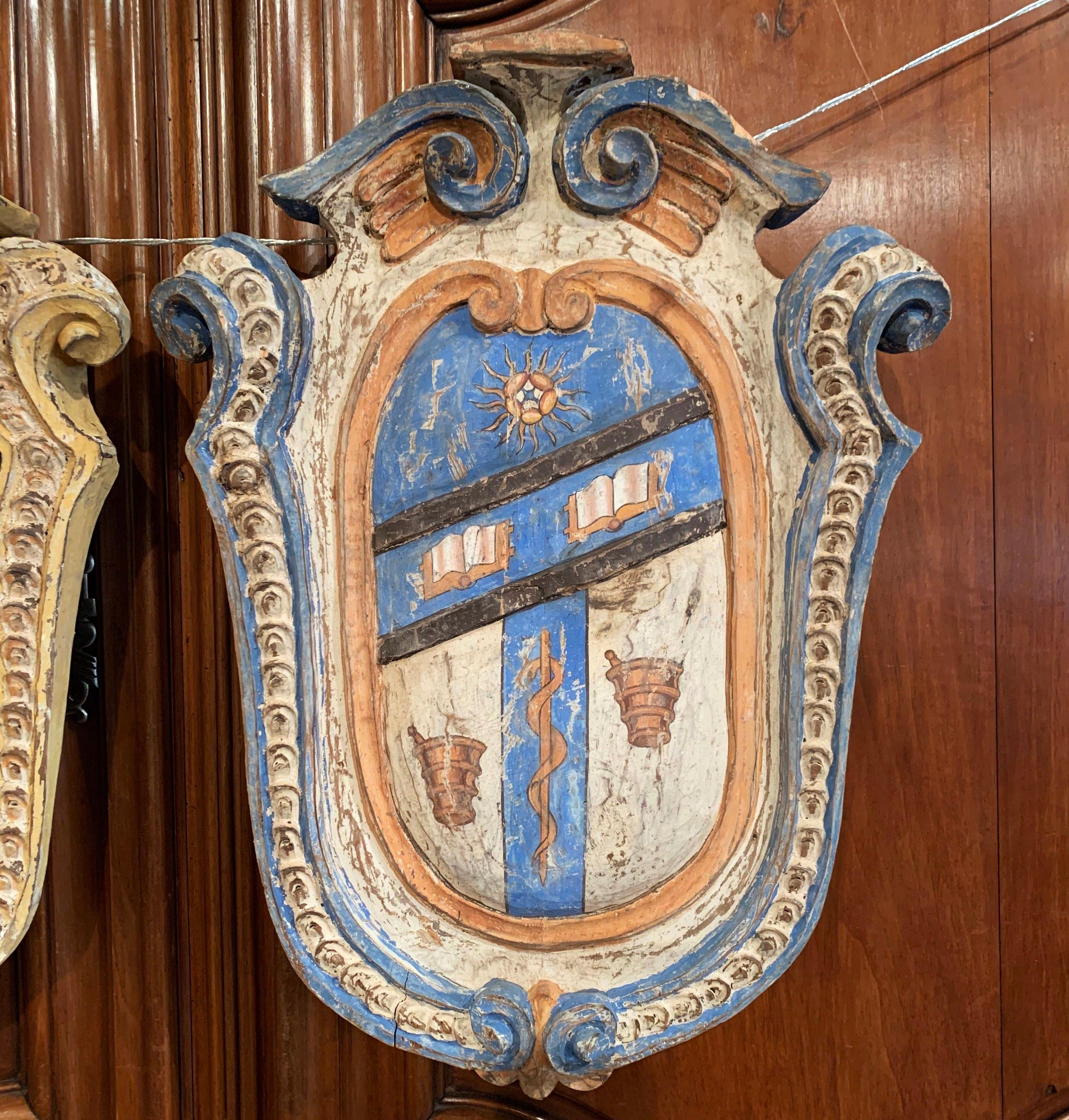 Embellish your study or library with this colorful pair of antique shields. Hand carved in France, circa 1920, each traditional crest features a central coat of arms decorated with hand painted family emblems including lion, book, mortar and