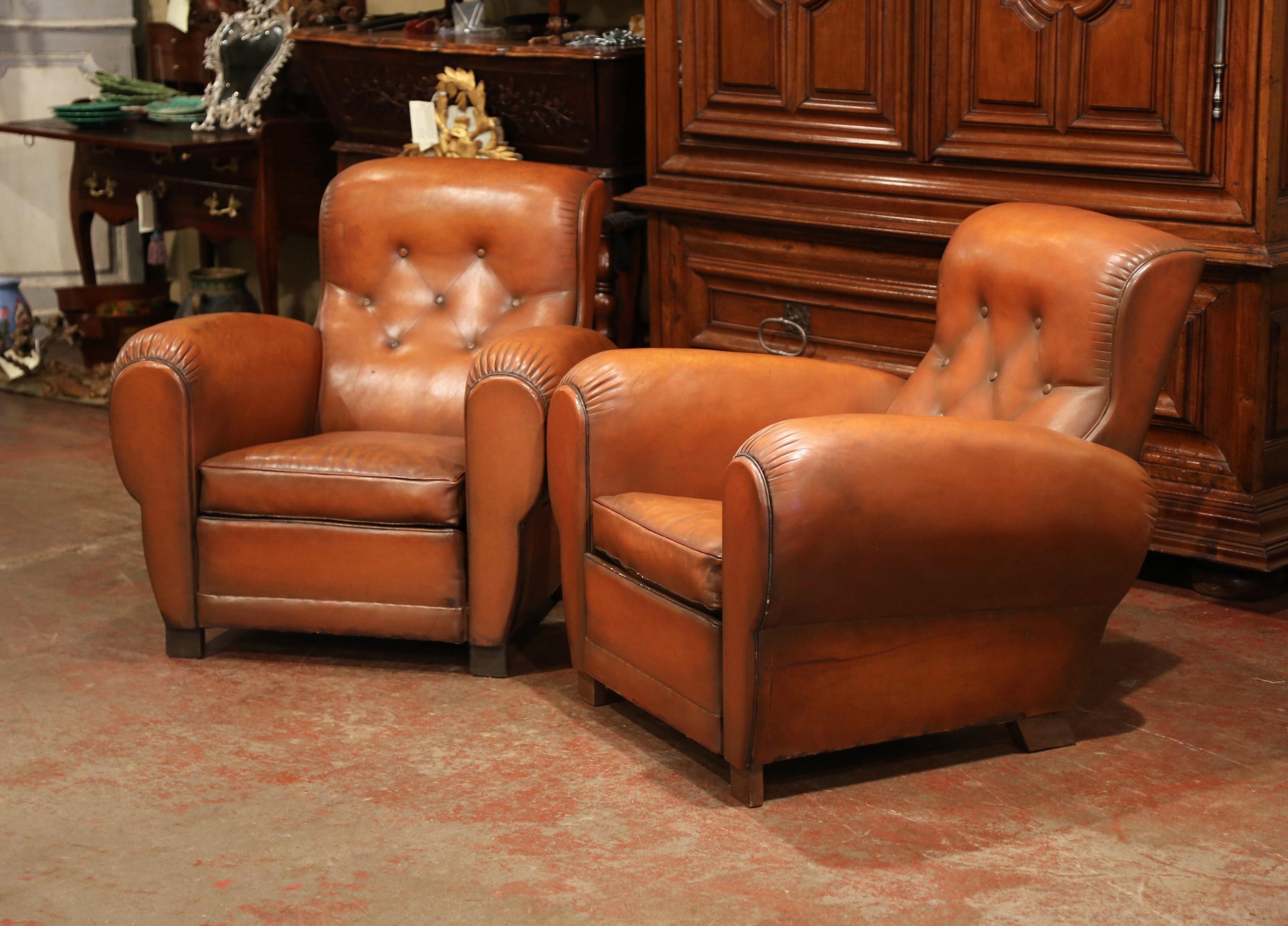 These cozy, antique Art Deco club chairs were crafted in France, circa 1920. The stately, masculine chairs feature wide, rounded armrests, a pitched back with ear shaped sides, and square wooden feet at the base. The comfortable armchairs are in