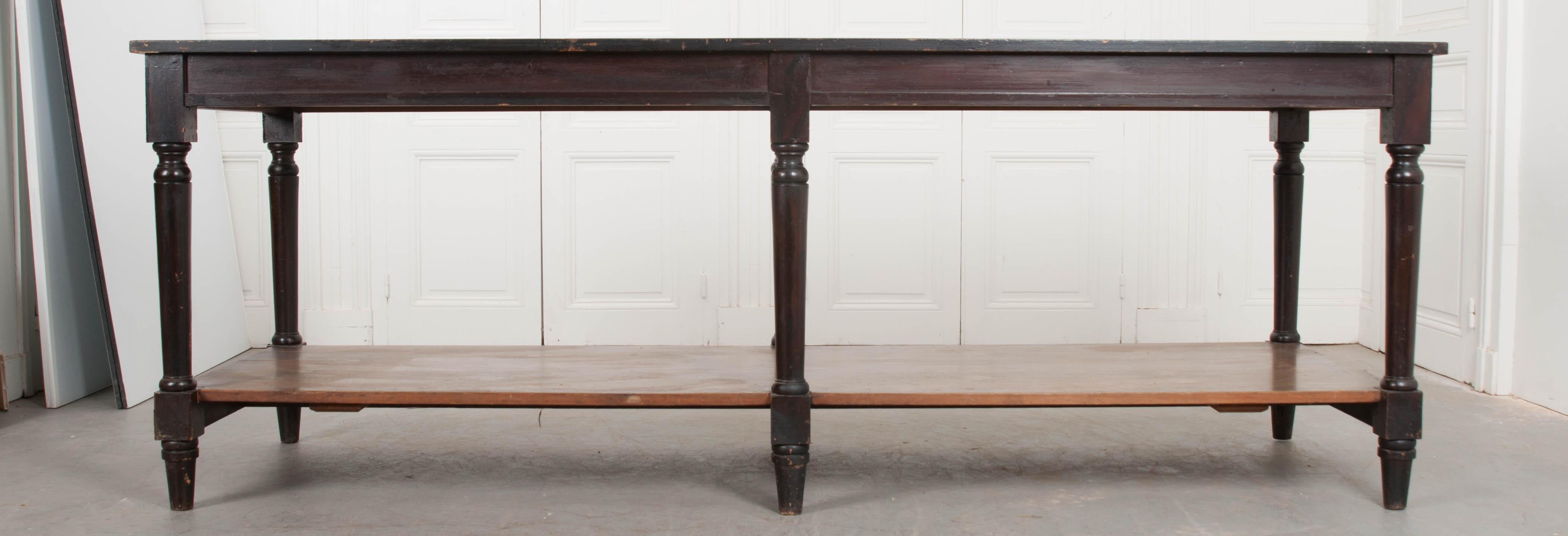 A gripping pair of dark drapery tables from 1900s France. The long tables both have tops that are painted black, slightly darker than the dark brown stain used to color the apron and legs. There is an undertier shelf that runs the length of each