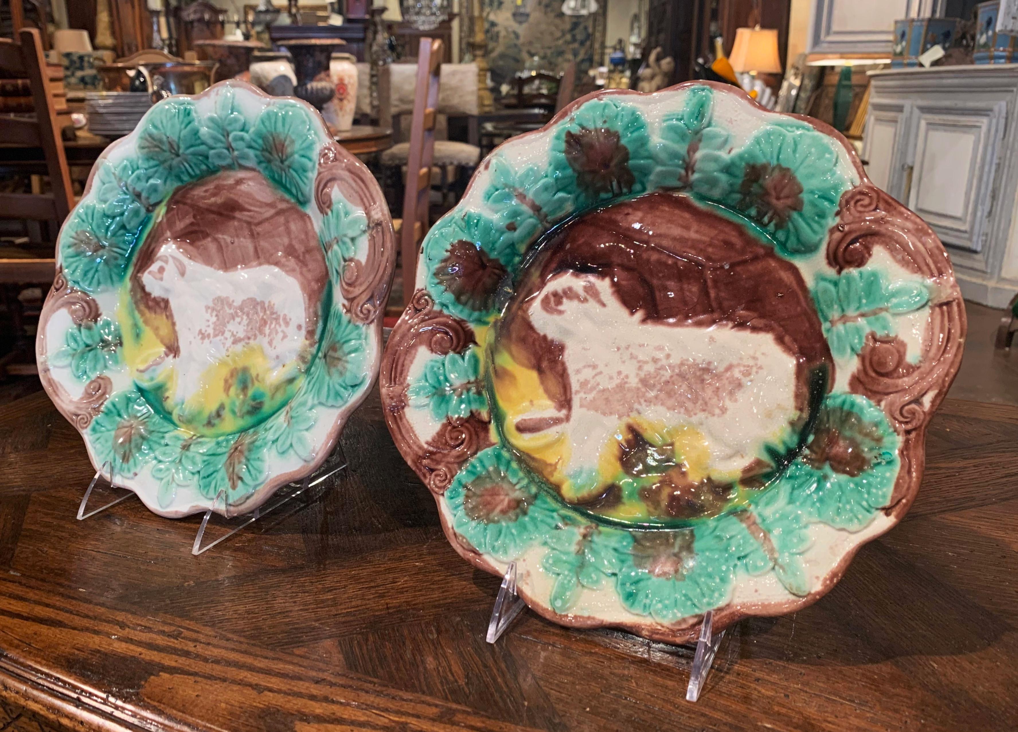 Decorate a kitchen wall with this colorful pair of antique ceramic platters, crafted in France circa 1930, each Majolica plate with a scalloped edge around the rim features leaf decor and embellished with a dog figure in relief in the center. The