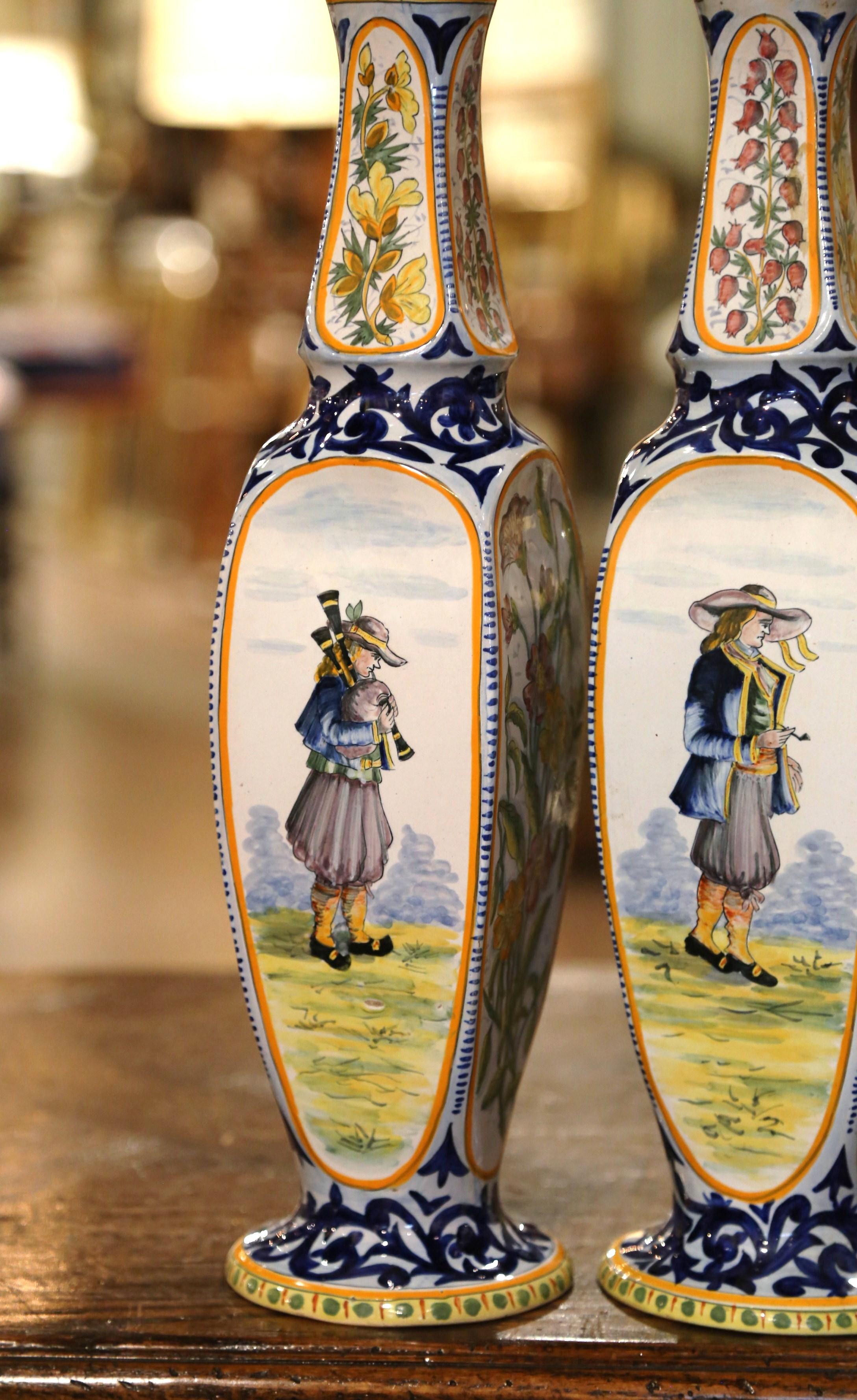 These elegant antique vases were crafted in Britanny France, circa 1920. The tall ceramic vases stand on a circular base over four flat sides and ending with a long neck. Each vessel features hand painted Breton people in traditional clothing,