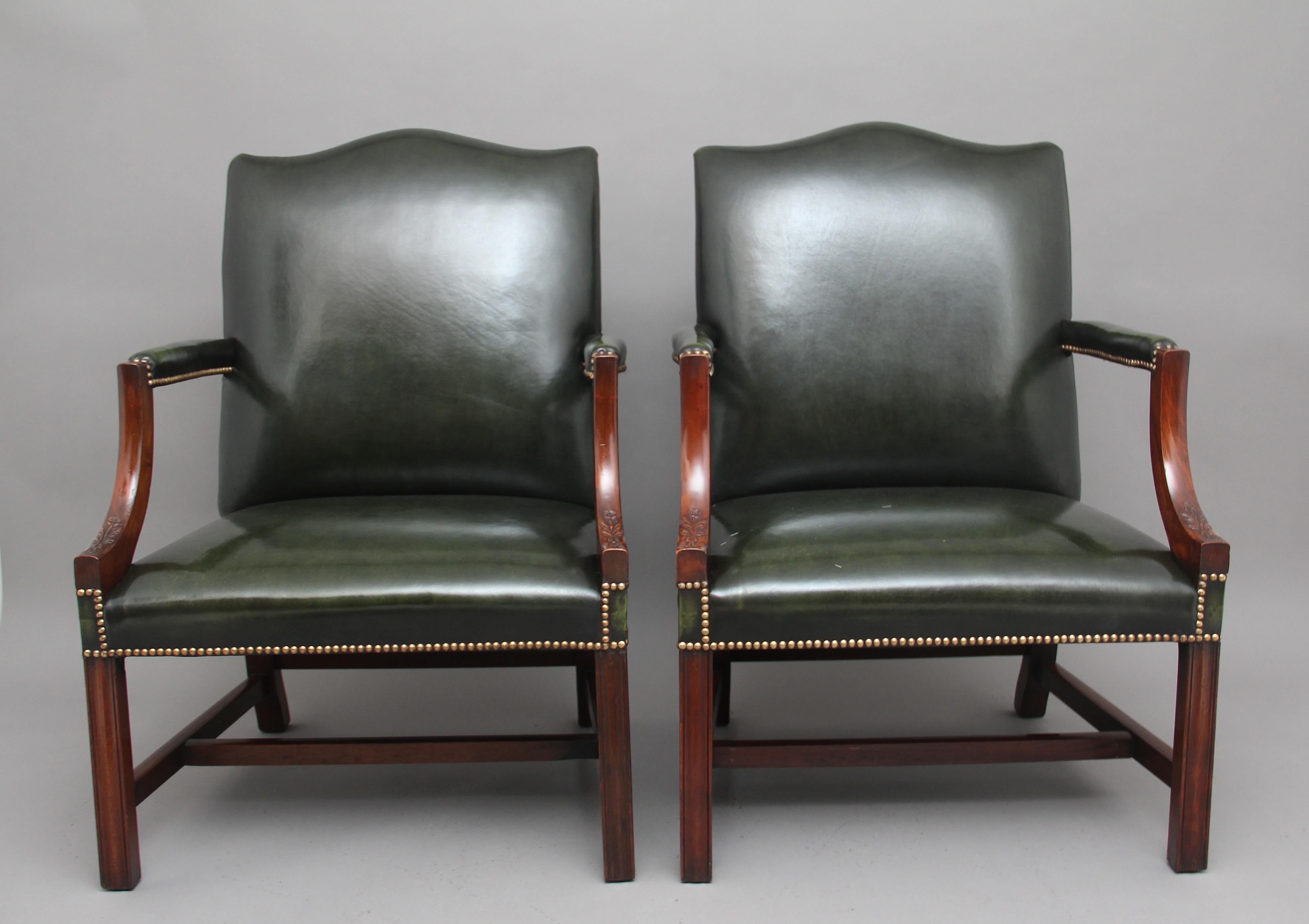 A pair of early 20th century mahogany Gainsborough / library open armchairs, upholstered in green leather with brass stud decoration, having padded arms and shaped arm supports carved with flowers, supported on moulded square legs united with