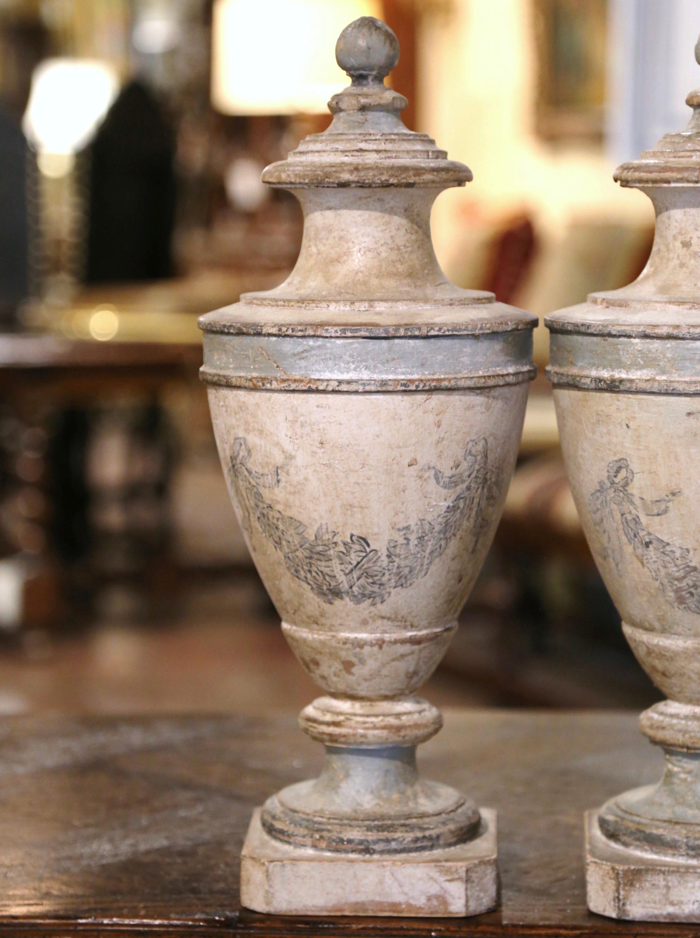 Decorate a mantel or a console with this elegant pair of antique urns. Crafted in Italy circa 1920, each tall vase is hand carved and decorated with a round finial; it features hand painted neoclassical swag and ribbon bow motifs on the main body.