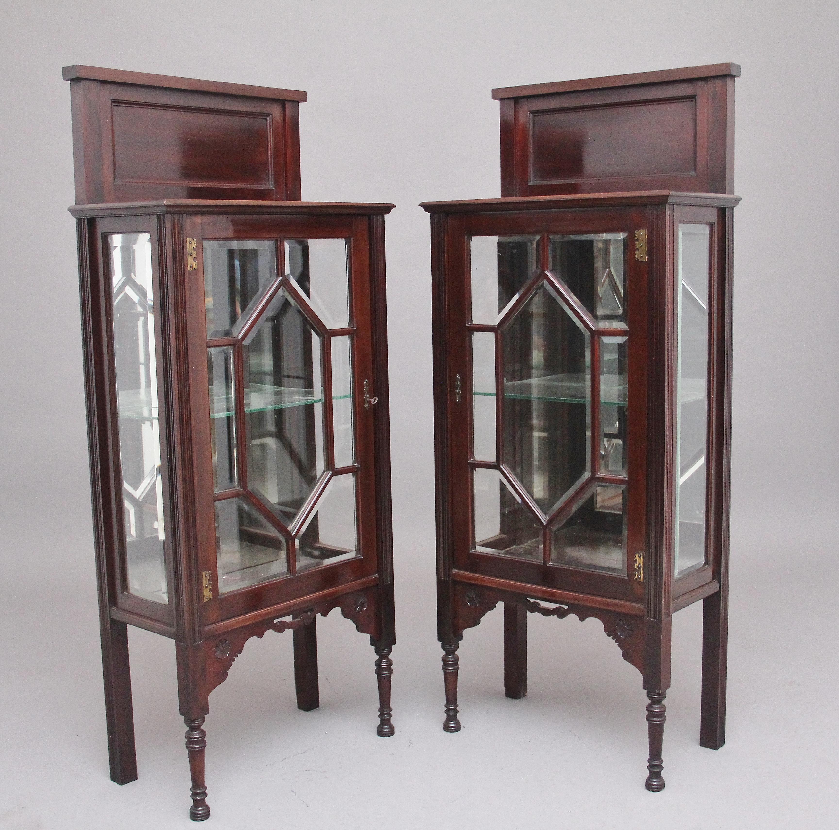 A pair of early 20th century mahogany display cabinets, the moulded edge top with a panelled back, astragal glazed door opening to reveal a mirrored back and a single glass shelf, having a decorative shaped and carved apron, supported on rear square