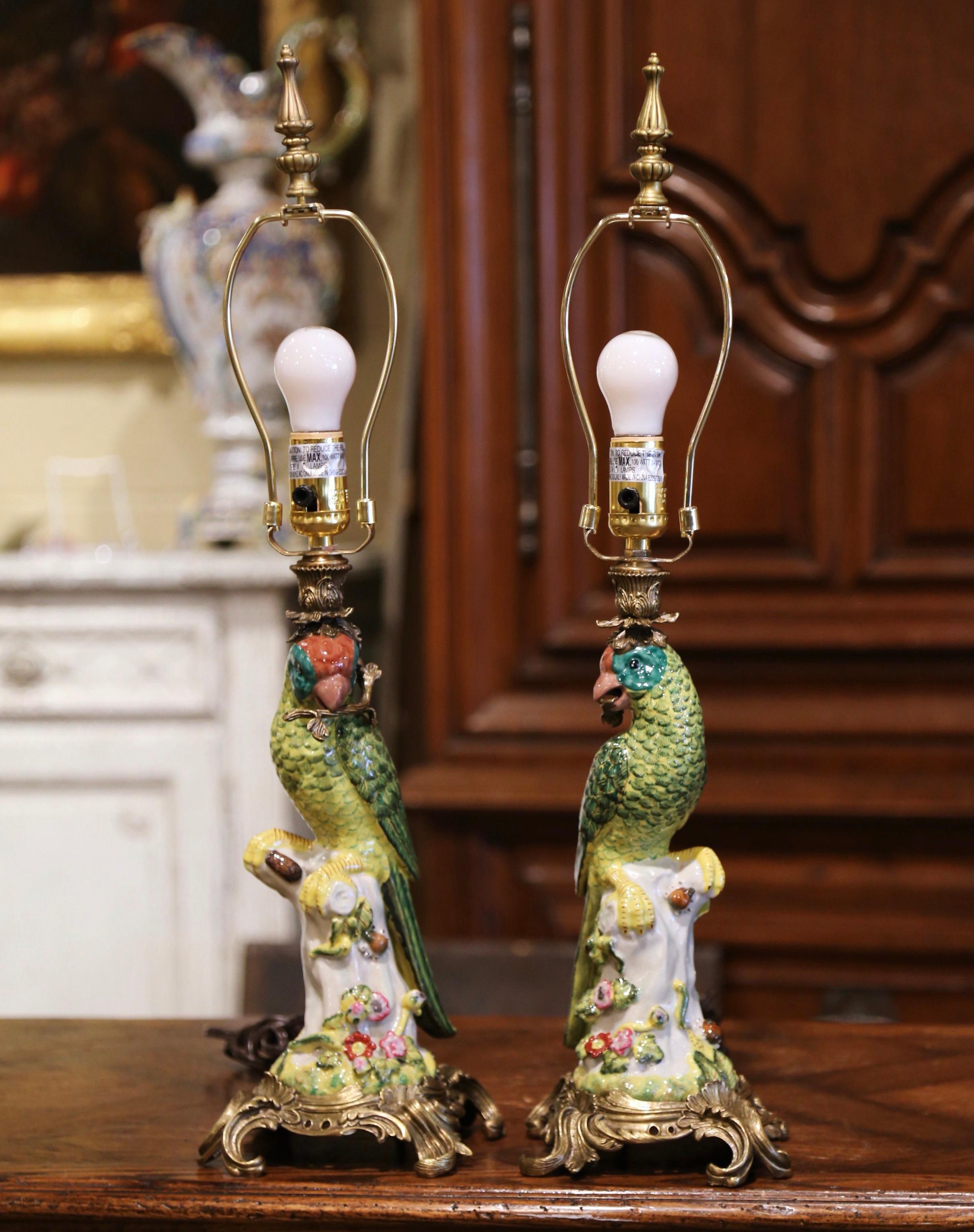 Decorate a bedroom or a den with this elegant pair of colorful antique lamps. Crafted in France circa 1920, each lamp stands on a bronze dore base decorated with leaf and shell motifs over foliage form scrolled feet. The hand painted bird sits on a