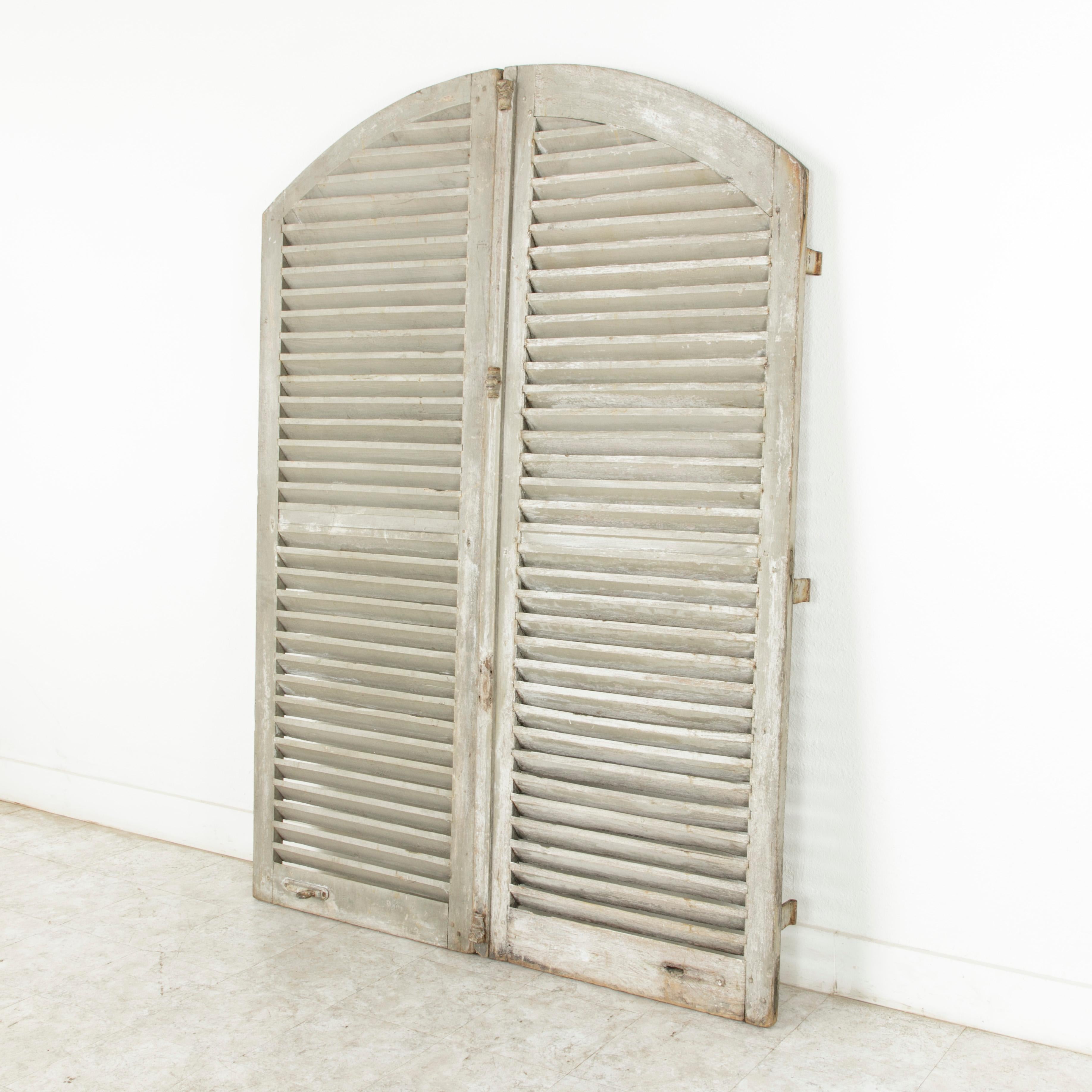 Standing at an impressive 82.5 inches in height, this pair of French shutters from the early 20th century features an arched top, slanted slats, and their original hardware. With a 61 inch total width, this tall pair of shutters painted in pale grey