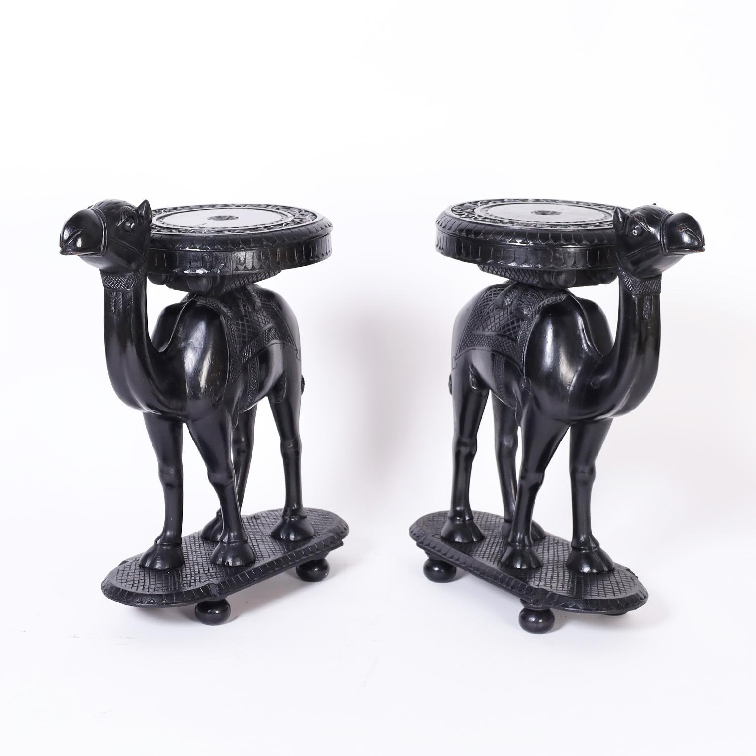 Pair of Anglo Indian camel form stands crafted in mahogany with an ebonized finish featuring round tops with floral carvings over expertly carved camels on bases with ball feet.