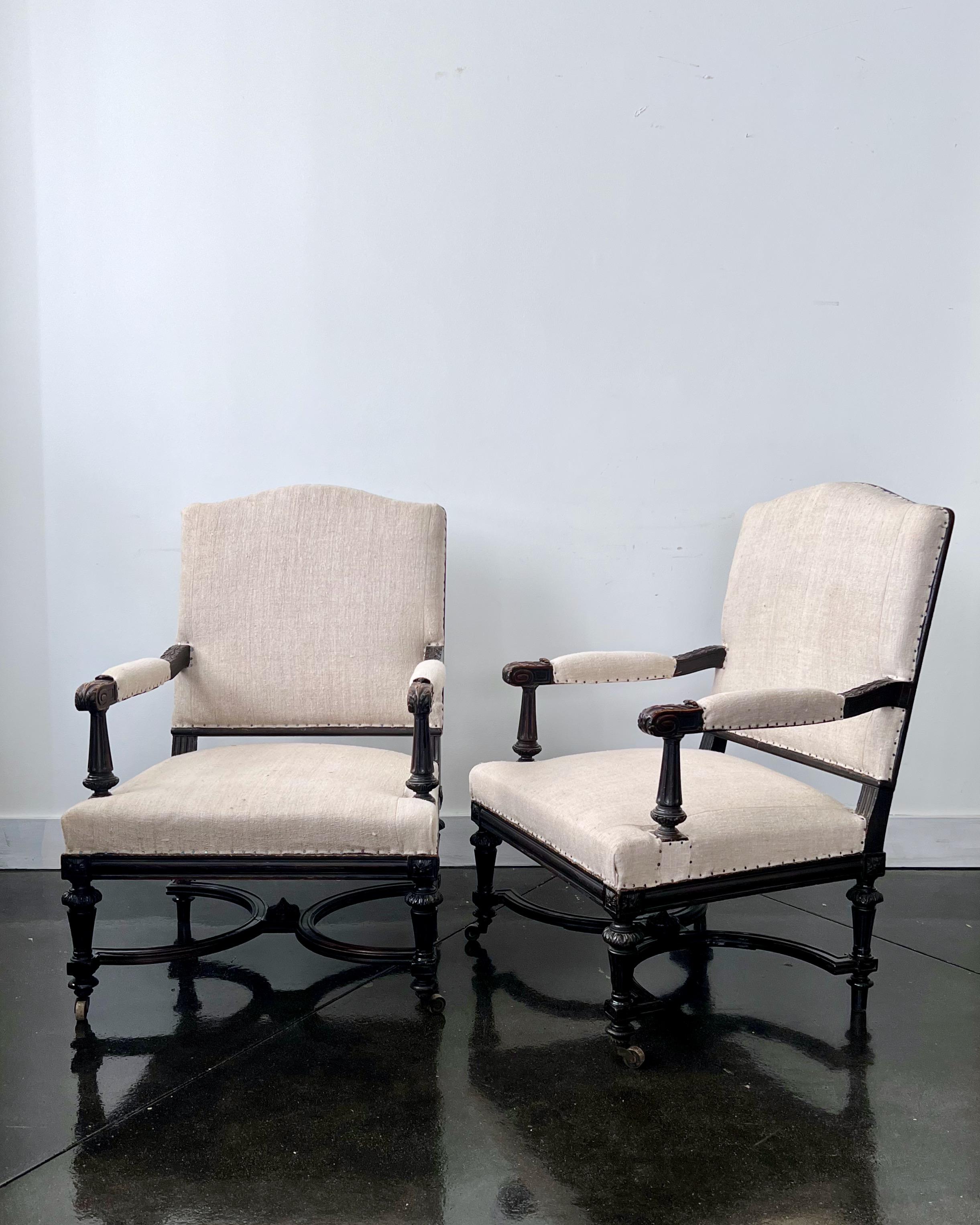 A handsome, very generous size  pair of ebonized French 19th century Napoleon III armchairs with richly carved armrests and raised on turned legs with casters on the front legs -and connected with cross finial decorated stretcher.
Upholstered with