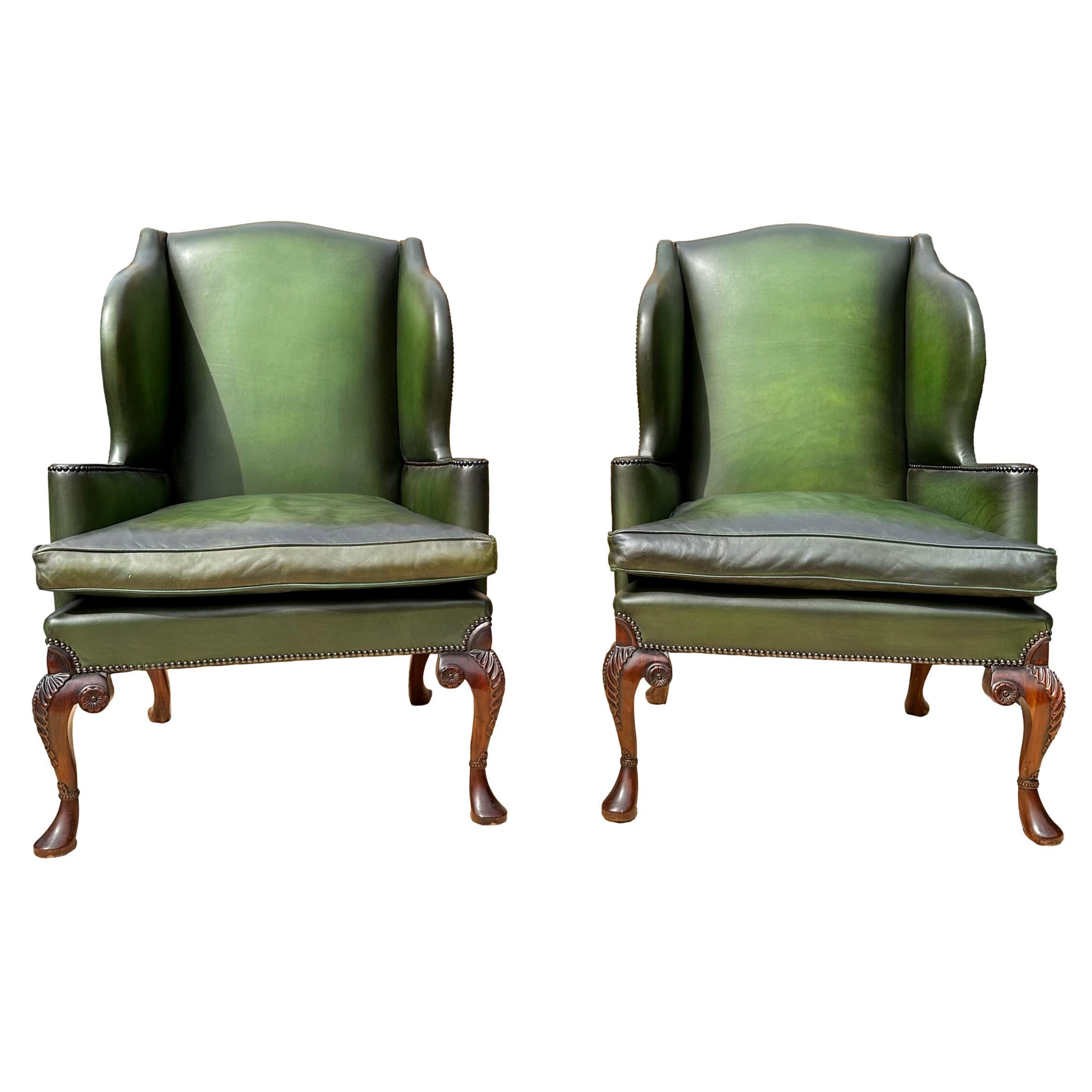 A Pair of Edwardian Green Leather Wing Back Chairs, beautifully proportioned with deep wings, the front legs carved with scrollwork and a Tudor rose, an acanthus leaf to the knees, and a carved cuff, finishing on a solid mahogany pad foot, with down