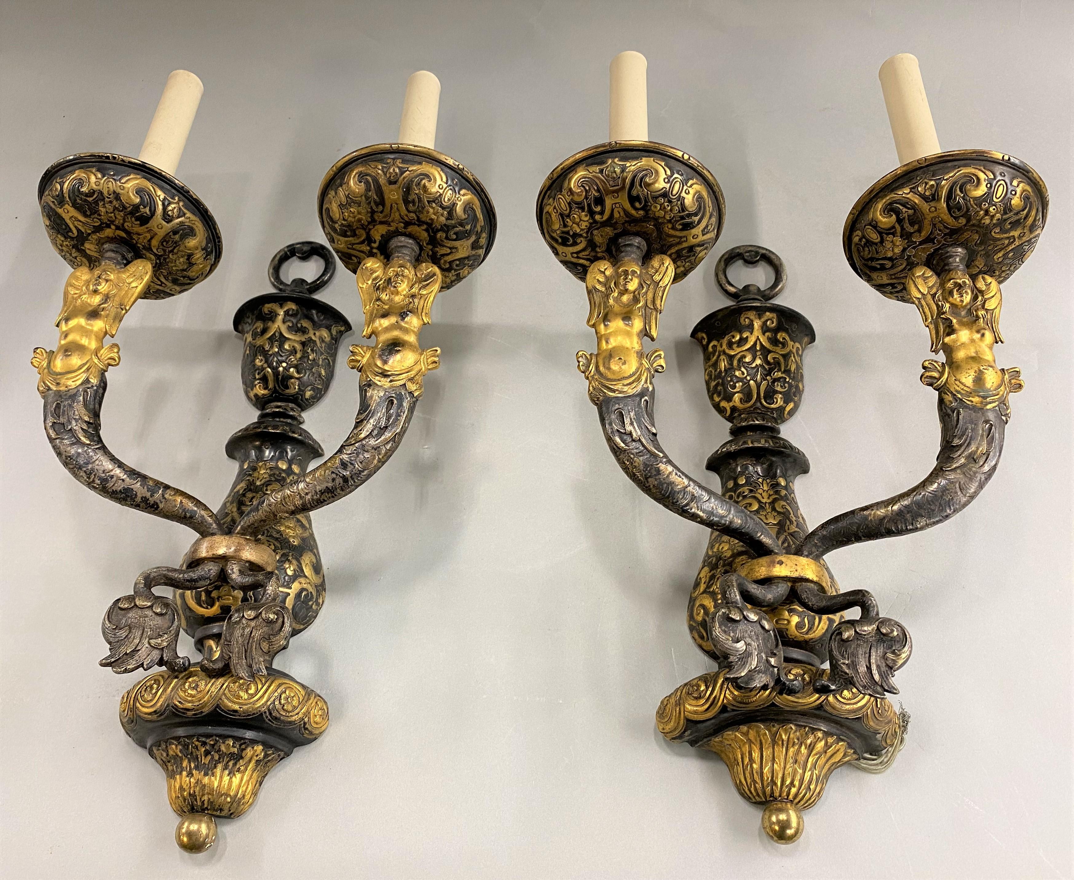 A beautiful pair of E.F. Caldwell two light scroll decorated bronze sconces with gilt figural arms showing angelic figures with mermaid tails, mounted on an urn form backplate. Each is stamped with inventory numbers on the back of the top loops. The