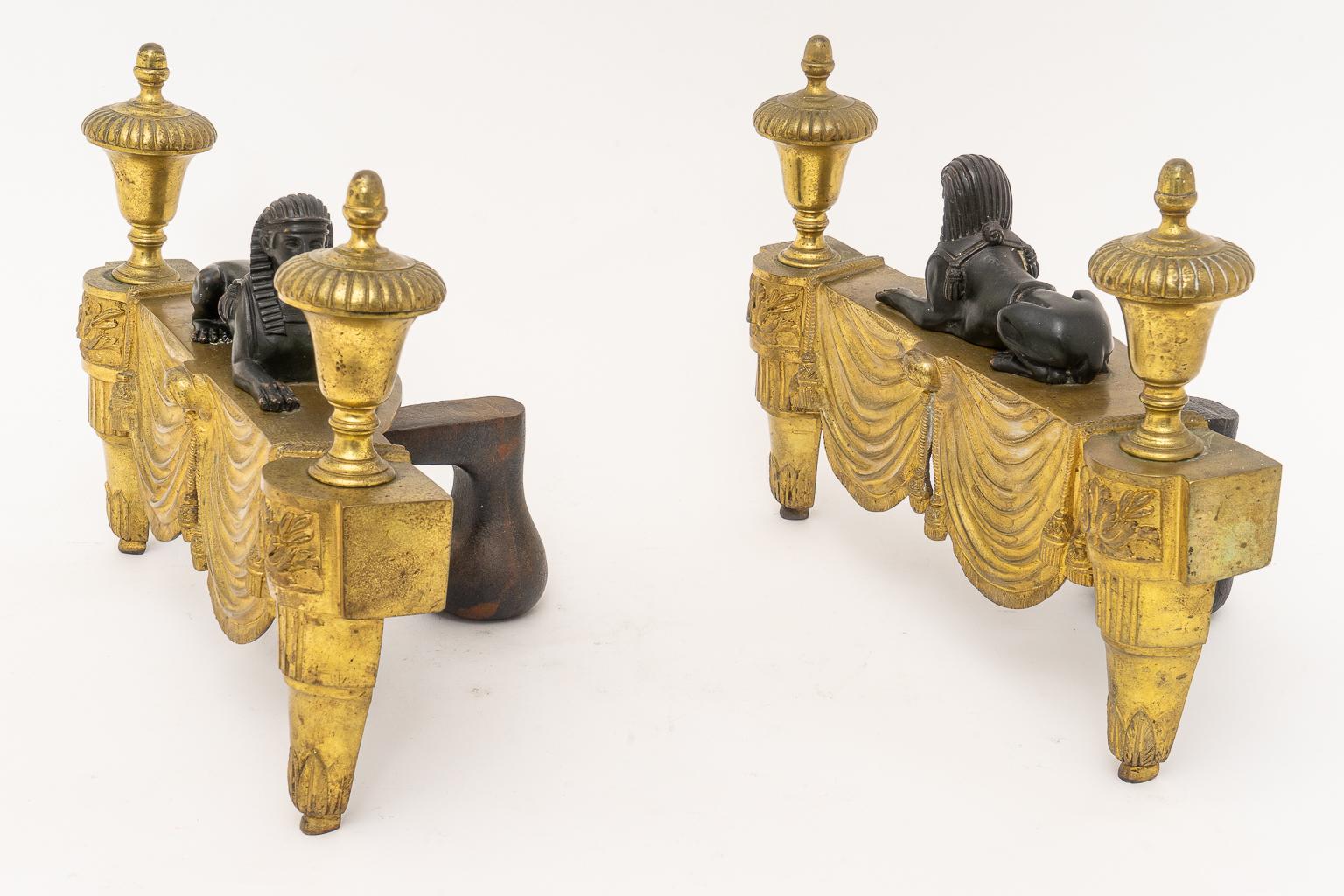 This stylish set of French, Egyptian Revival chenets were acquired from a Palm Beach estate. The pieces are fabricated in gilt bronze and patinated bronze. The dark sphinx contrast handsomely against the gilt bronze base that is detailed with swaged